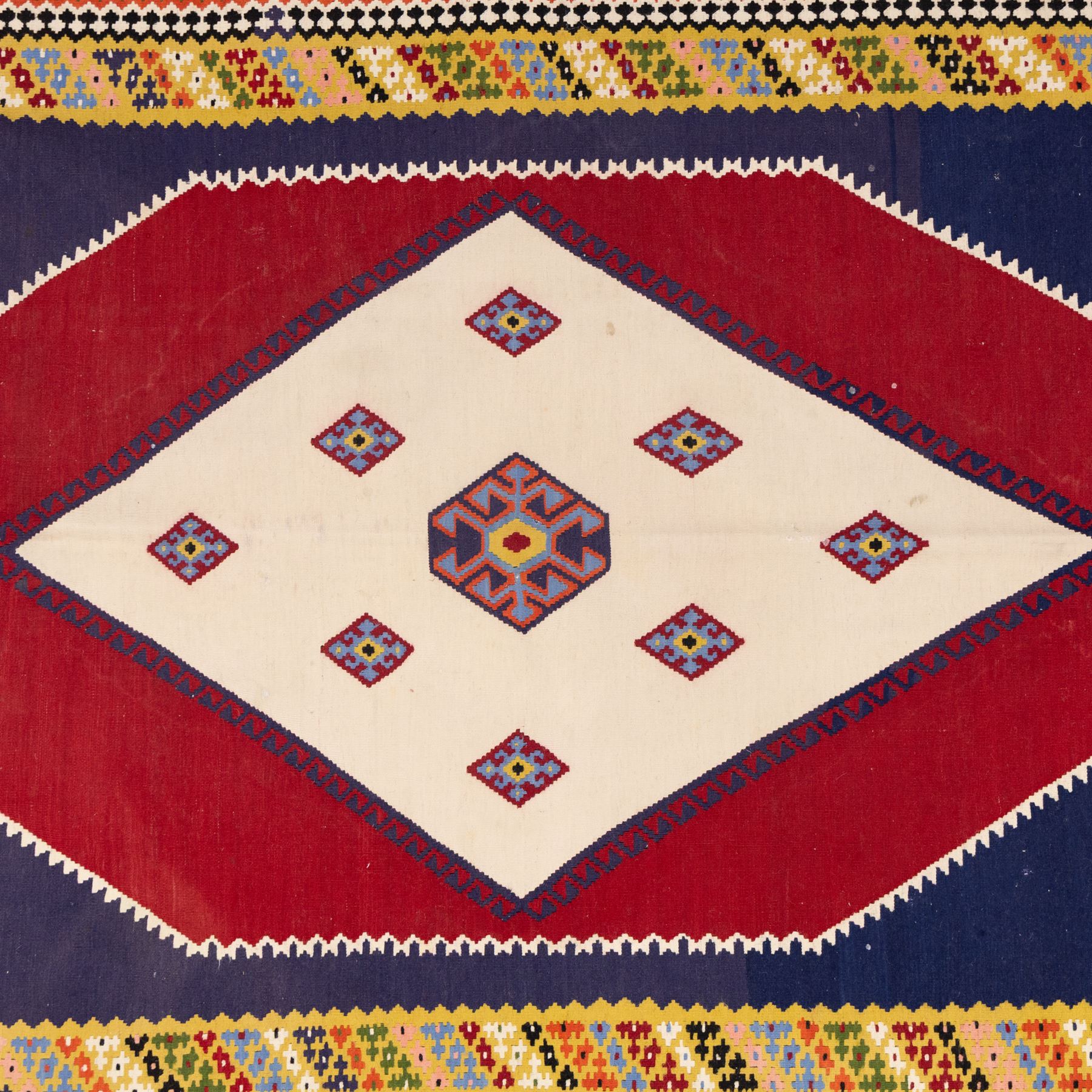 South West Persian Qashqai kilim, woven in flatweave with tones of indigo blue, madder red, yellow, turquoise, and green, with a bold central diamond medallion on an ivory ground, enclosing smaller lozenge motifs and flanked by deep madder red panels, field framed by a broad indigo border with polychrome geometric devices and serrated motifs 