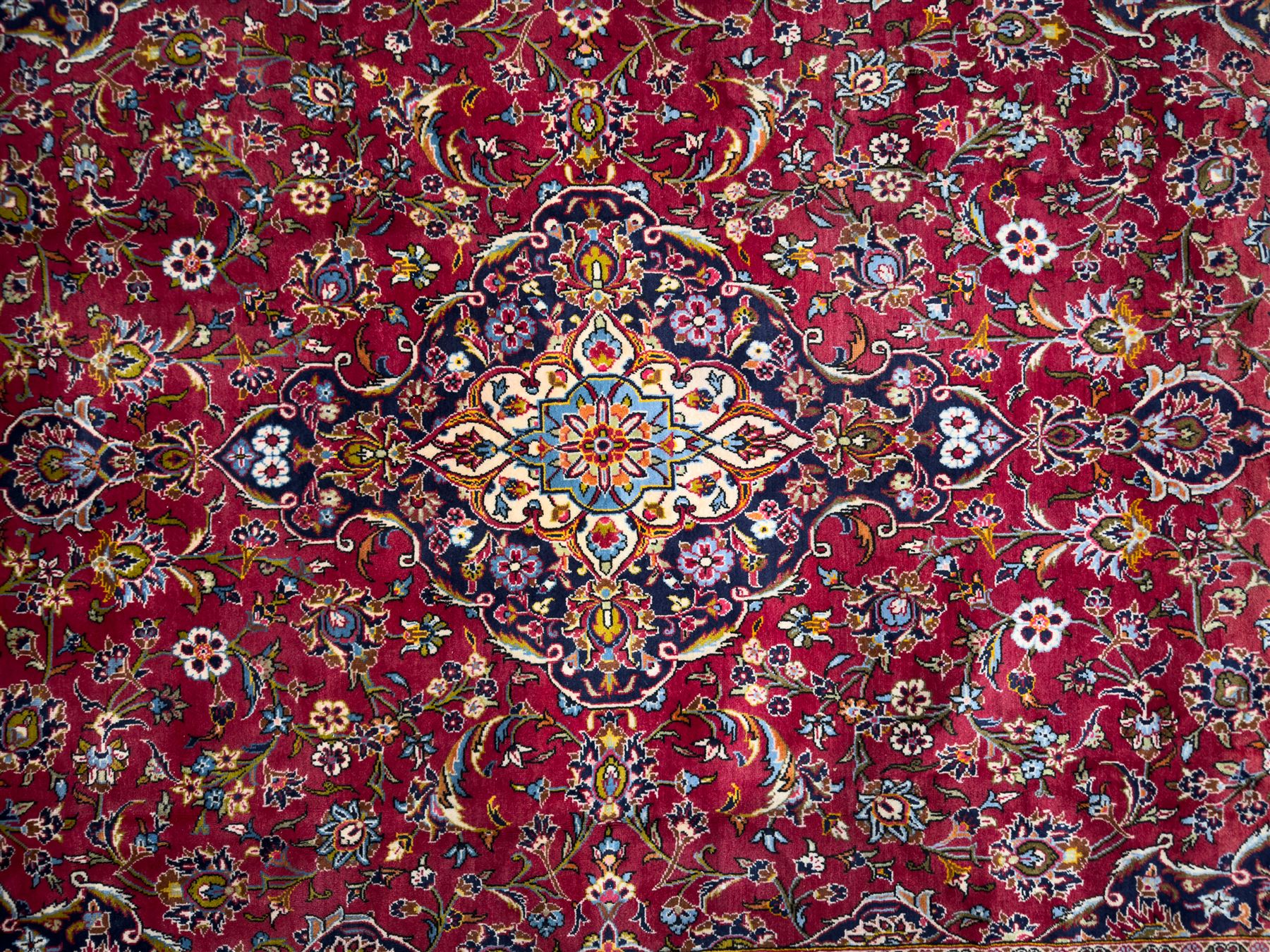 Central Persian Kashan hand-knotted carpet, the madder red field with central lobed medallion in indigo and ivory, enclosing scrolling arabesques and flowerheads, framed by palmette spandrels and an indigo border of rosettes and lotus forms between multiple floral guard stripes, from the region of Kashan