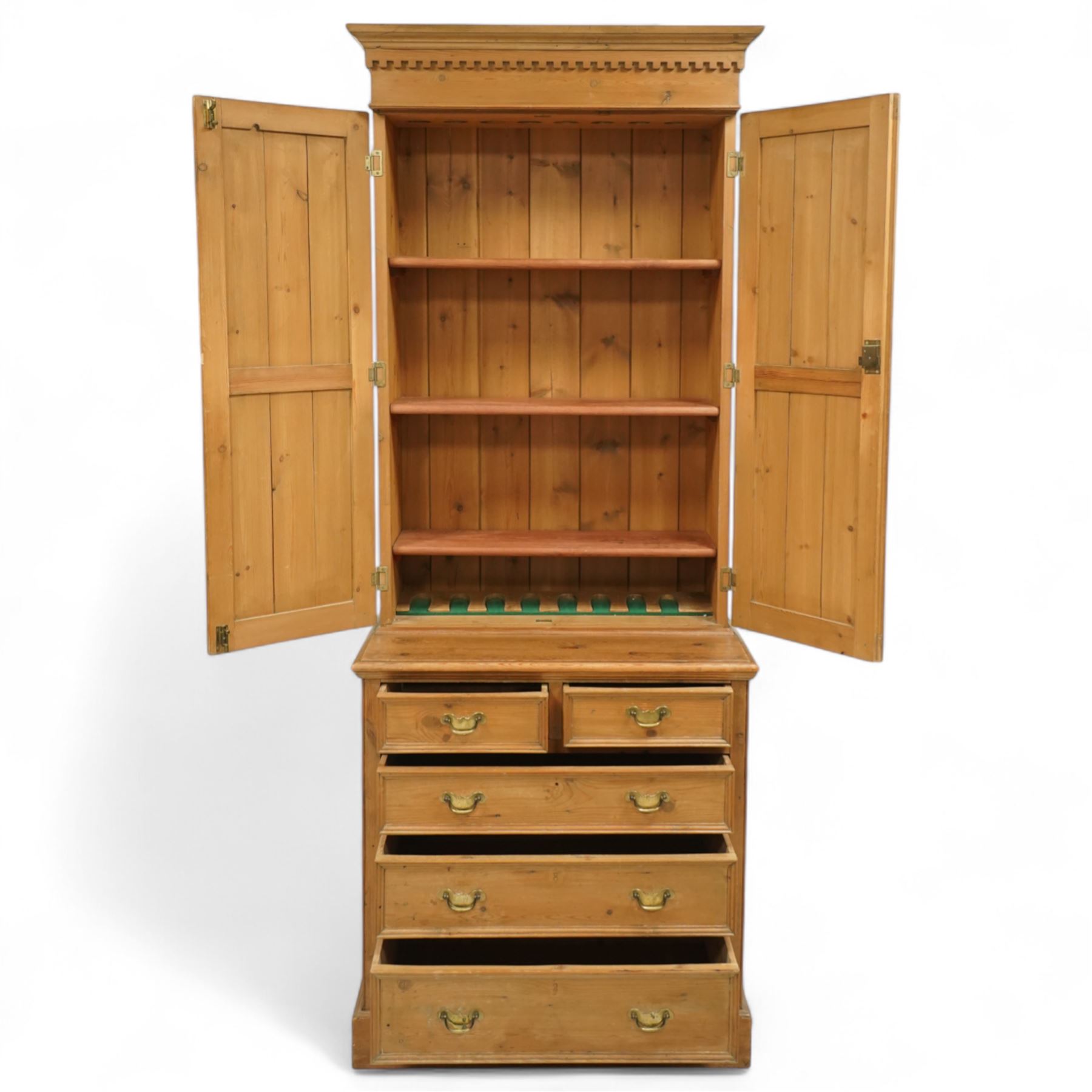 Early 20th-century pine gun cabinet with later fitted shelves, of two-part construction, upper cupboard moulded and dentil cornice above two panelled doors revealing three removable shelves and circular recesses for gun barrels at the top, lower section with two short and three long graduated drawers with brass swan neck handles on plinth base 