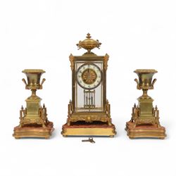 French- Art-deco Edwardian 8-day four-glass clock garniture, with etched glass panels, cast brass decoration to the corners of the case and splayed feet representing cloven hoofs to the base, case surmounted by a large oval urn on a tiered brass platform, two part dial with a recessed brass centre comprising of pierced radial decoration, with steel spade hands and Arabic numerals to the chapter, rack striking movement with a deadbeat Brocot escapement, striking the hours and half-hours on a bell. With a visible twin file mercury pendulum. Garni comprising two large twin handled urns on rectangular pedestals raised on conforming feet representing cloven hoofs, Clock and garni presented on three shaped wooden gesso bases with velvet pads. With key.