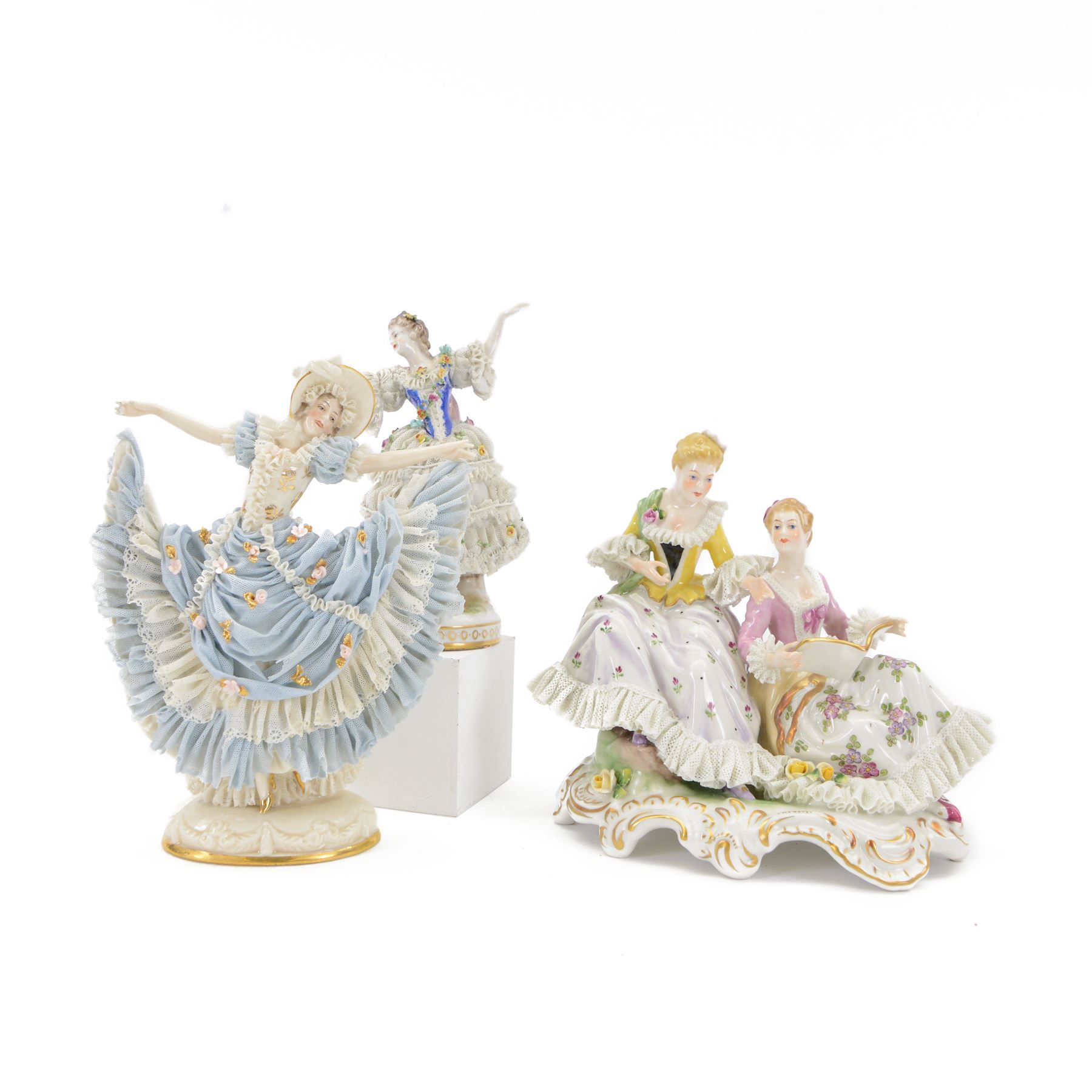 Three Continental lace figures, comprising a figure group modelled as two seated female figures reading, with lace trimmed gowns, H19.5cm, the other two examples modelled female figure with arms outstretched, the lace gowns encrusted with flowers, taller example H21cm