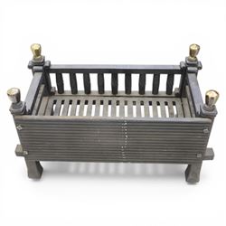 Arts and Crafts cast iron fire grate, rectangular backplate with plain upper rail over a slatted front of cylindrical tapering bars, flanked by square section uprights with fluted detail and scroll terminals, raised on stepped feet with shaped brass roundel mounts and brass finials to the corner posts