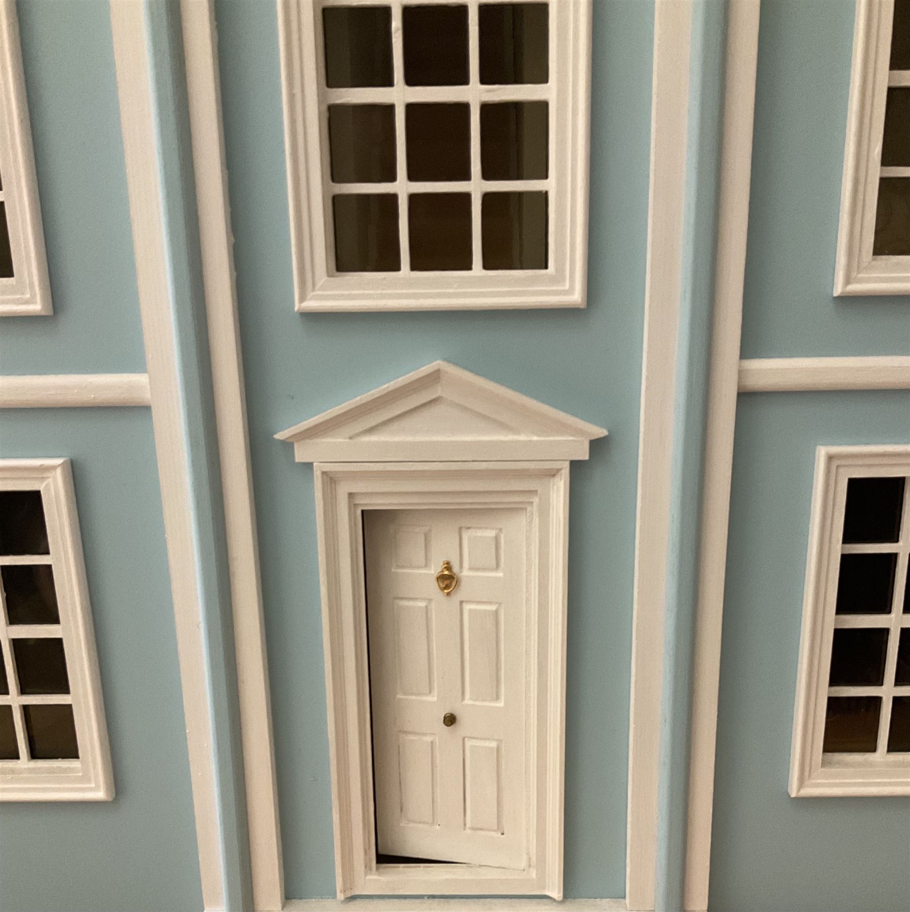 Georgian style wooden double fronted three-storey dolls house with pale blue stucco finish under a faux tiled roof with two chimney stacks, the double hinged front opening to reveal six fully decorated rooms with floor coverings, central staircase, porcelain fitted bathroom, kitchen with deep white sink and Aga cooker, electric lighting with adaptor, fully furnished with French style and other furniture, grand piano etc, and comprehensive range of accessories H82cm W61cm D31cm