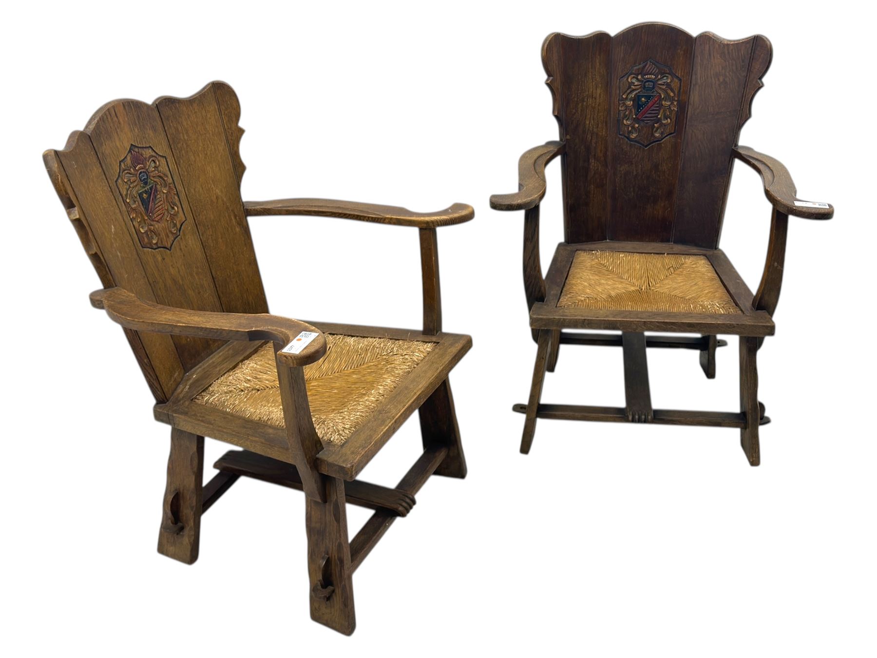 Pair of Arts and Crafts oak armchairs each with shaped backrest centred by polychrome painted crest, rush seat over straight square legs united by shaped central stretcher