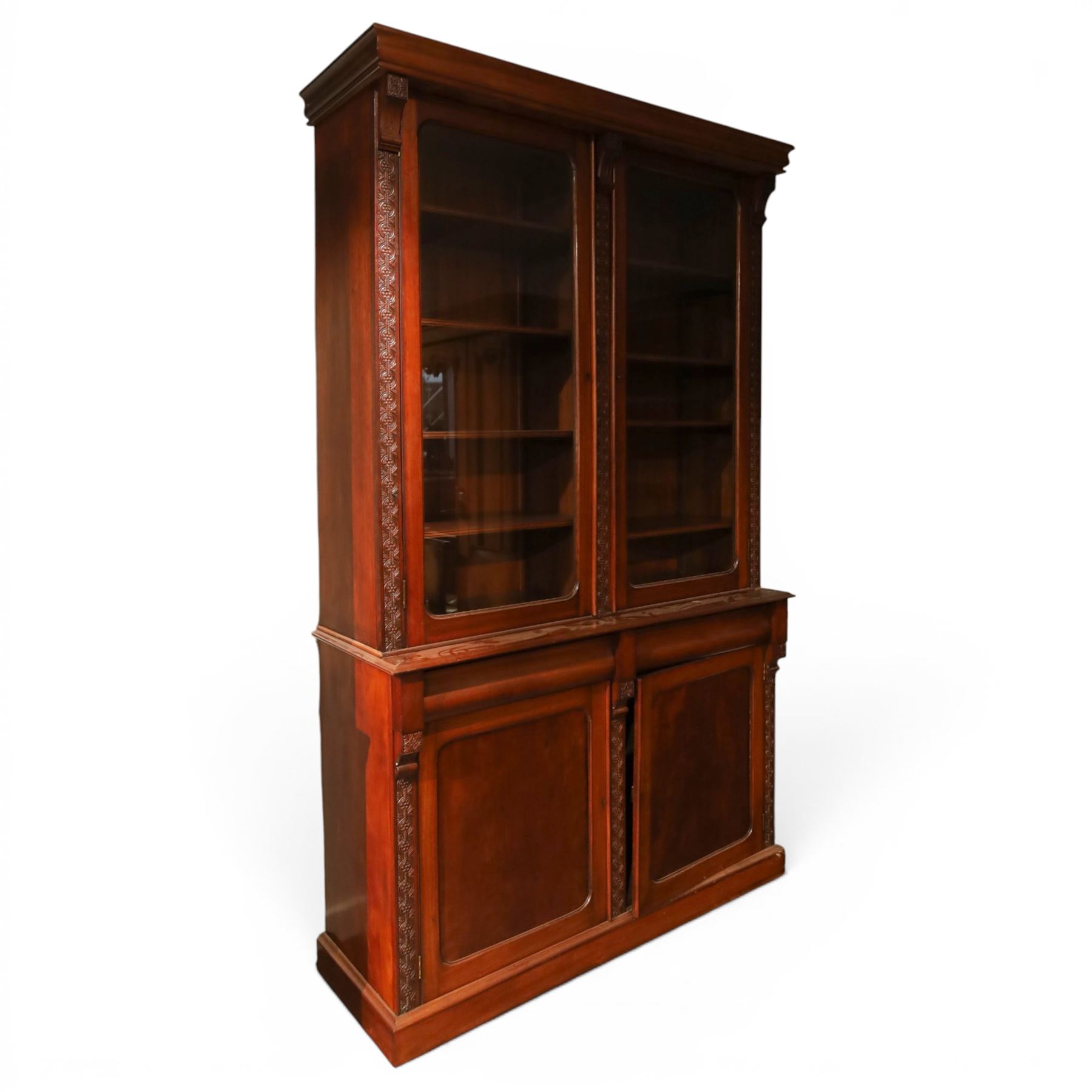 Victorian carved mahogany enclosed bookcase on cupboard, projecting moulded cornice over twin glazed doors enclosing adjustable shelves, flanked by foliate carved uprights, the lower section fitted with two cushion moulded drawers over twin panelled cupboard doors, divided and flanked by conforming carved uprights, on plinth base