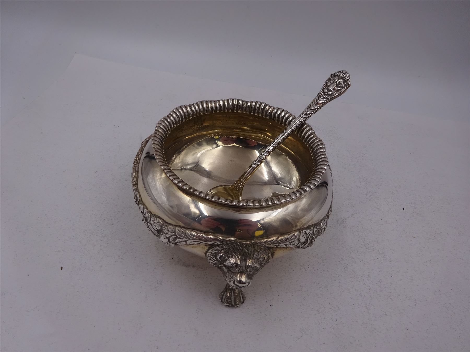 Early 20th century silver open sucrier, of circular form, with fluted gadrooned rim and decorated in relief with floral swags, united by three lion mask mounts and upon three paw feet, hallmarked Z Barraclough & Sons, London 1912, H6cm, D11cm, with similar spoon with lion mask finial, hallmarked London 1912, maker's mark indistinct