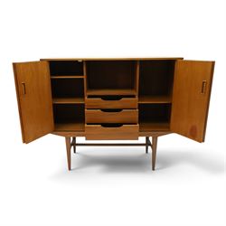 Alfred Cox - mid-20th century teak cabinet, rectangular top over two bifold doors with inset handles, opening to reveal three central graduating drawers surrounded by assorted shelves, raised on tapering supports united by H-stretcher, stamped to inside of drawer