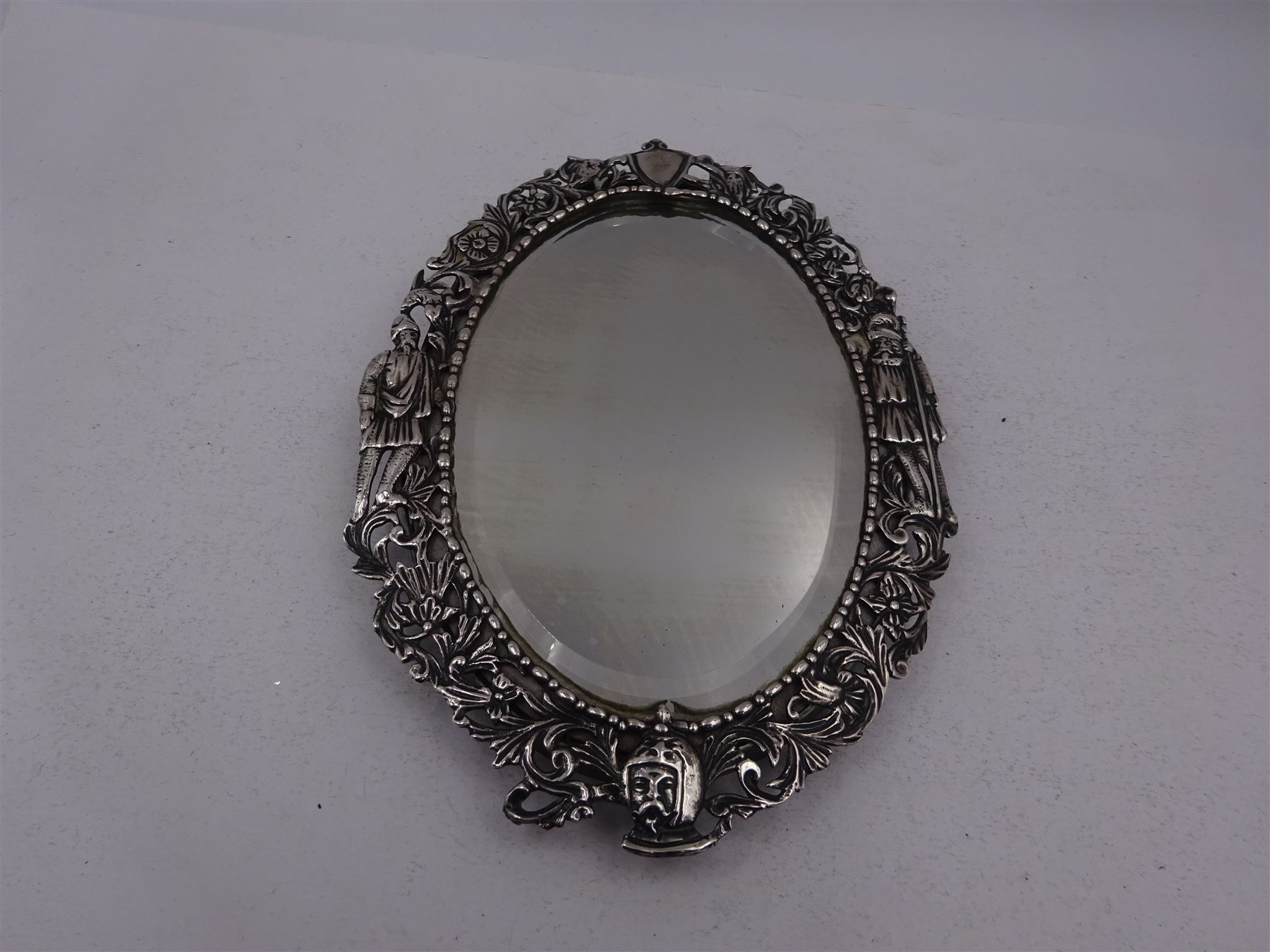 Continental silver miniature dressing table/wall mirror, of oval form with bevelled mirror plate, surrounded by a bead and dart rim, with pierced scrolling floral border with a standing cavalier to either side and the bust of a cavalier to the base, H17.7cm