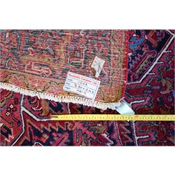 North West Persian Heriz hand-knotted carpet, brick-red field with a large central angular star medallion with pendant motifs, surrounded by stylized geometric palmettes, serrated leaves and tribal devices, ivory corner spandrels similarly decorated, field enclosed by a dark indigo main border of stylized rosettes and angular vine motifs between multiple guard stripes, with cotton fringe ends  