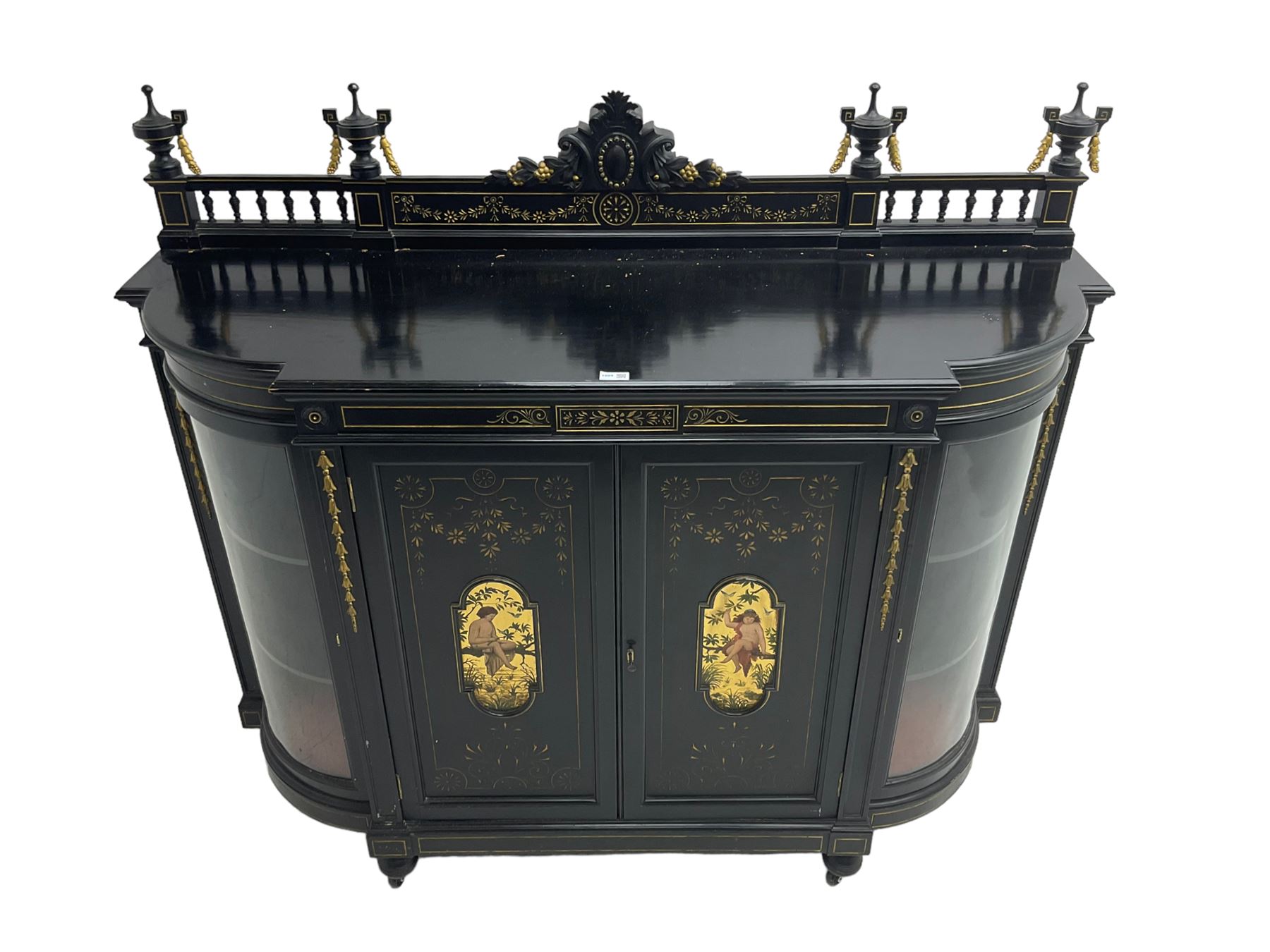 Victorian ebonised credenza side cabinet, raised balustrade back with urn finials and a central carved cartouche, carved and gilded with floral garland decoration, central doors with gilt panels painted with figures sitting over a lake enclosing two shelves, flanked by applied bellflower detail and glazed bow-front side doors, raised on ring turned feet with castors