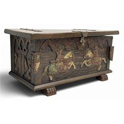 Mid-20th century carved hardwood chest, the rectangular hinged top with decorative strap hinges, the front and sides deeply carved with medieval-style jousting knights, heraldic shields, and architectural elements, raised on shaped block feet