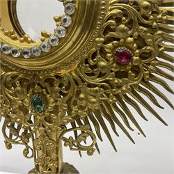 20th century gilt metal monstrance, in the Gothic revival style, with glazed aperture to centre surrounded with clear paste stones, in a pierced floral and scroll border set with coloured paste stones, within a sunburst surround, on a paste set knopped stem and similar foliate foot, H59.3cm