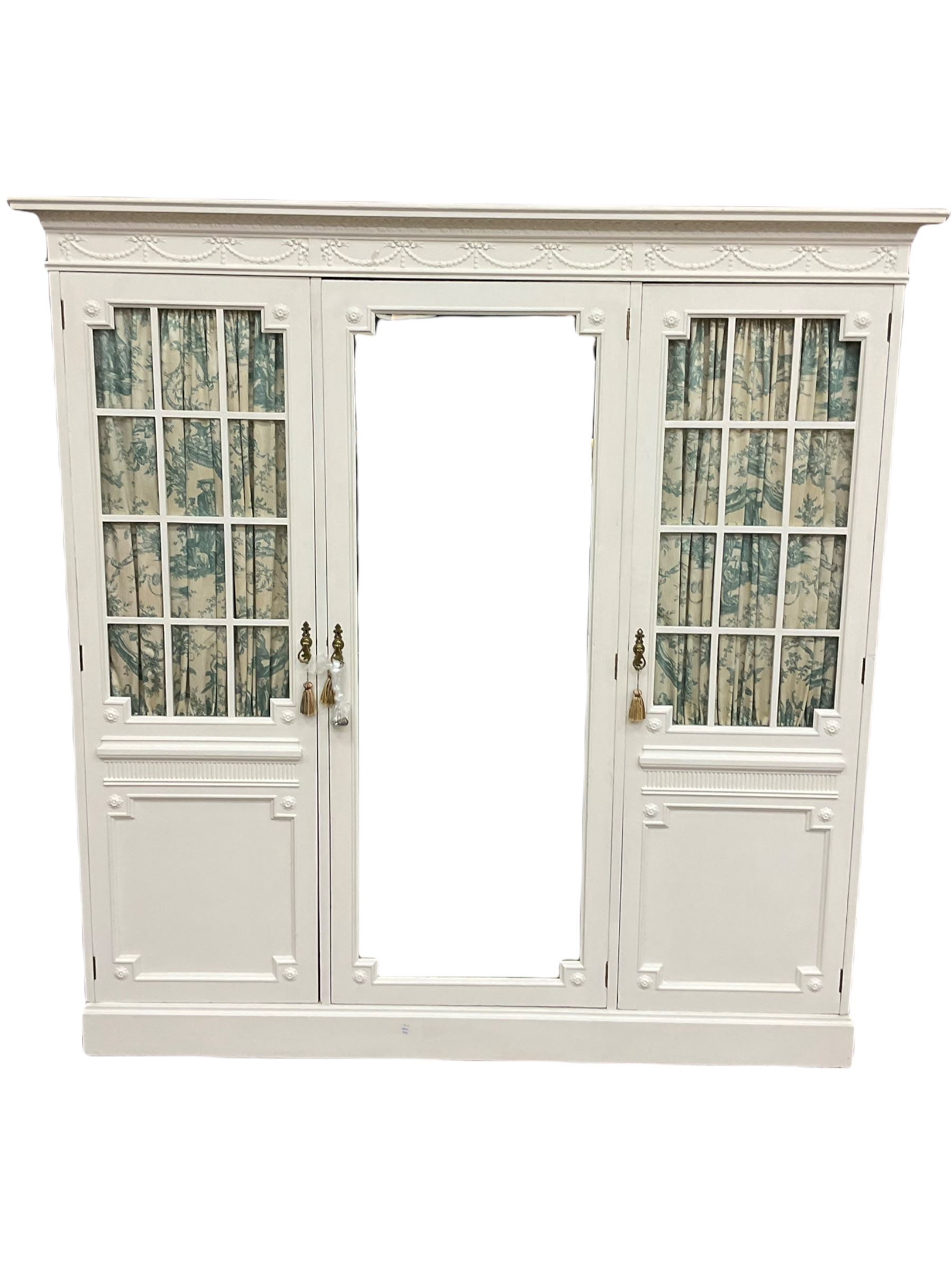 Maple and Co - Edwardian Hepplewhite design white painted triple wardrobe, foliate carved projecting cornice over ribbon and husk swag decorated frieze, two curtain lined astragal glazed doors and centre full mirror door, fitted mahogany interior including slides and drawers, plinth base, label to top of central door