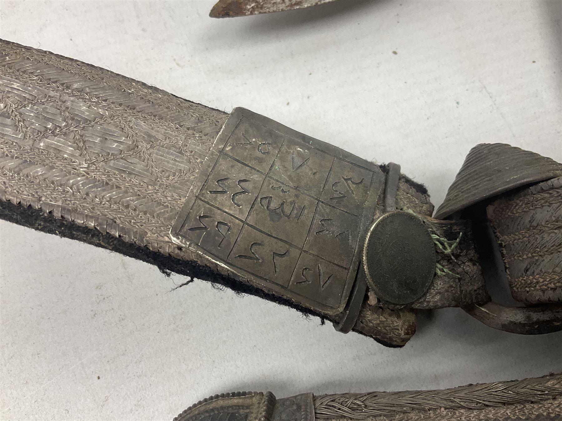 Omani Khanjar dagger, the 19cm curved blade with pronounced medial rib to either side, waisted hilt with dome shaped pommel and silver mounted horn grip decorated with an embossed band; in an intricately decorated silver scabbard with silver rings woven into the belt mount and fitted with original coin mounted silver thread-work belt with additional side knife in sheath verso L34cm overall, belt length 81cm; together with a Nepalese kukri in leather scabbard with one skinning knife (2)