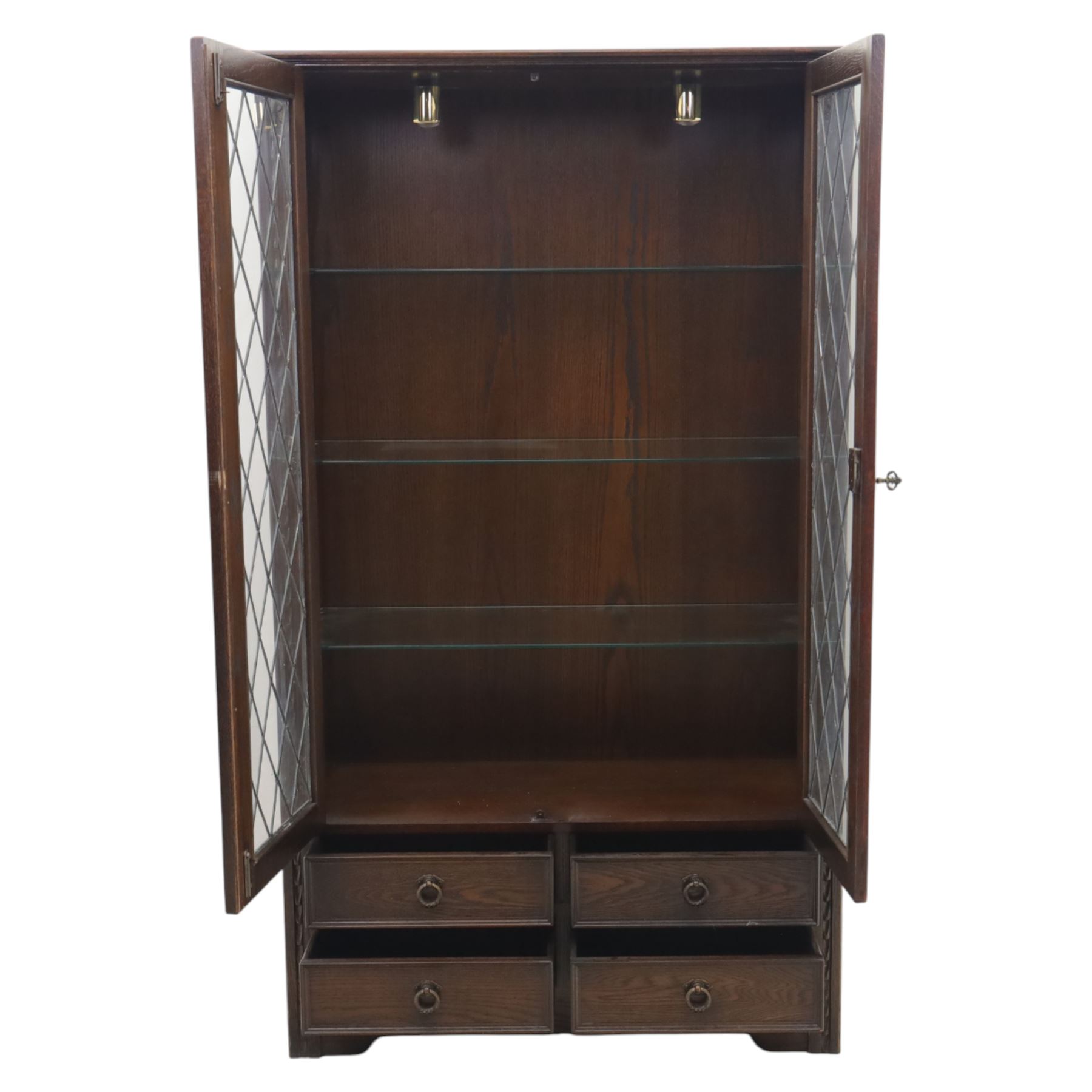 Mid 20th century oak display cabinet, the moulded rectangular cornice above a pair of leaded glazed doors with diamond lattice glazing, enclosing adjustable shelving, the lower section fitted with four short drawers with brass ring handles, flanked by chamfered uprights with carved leaf detail