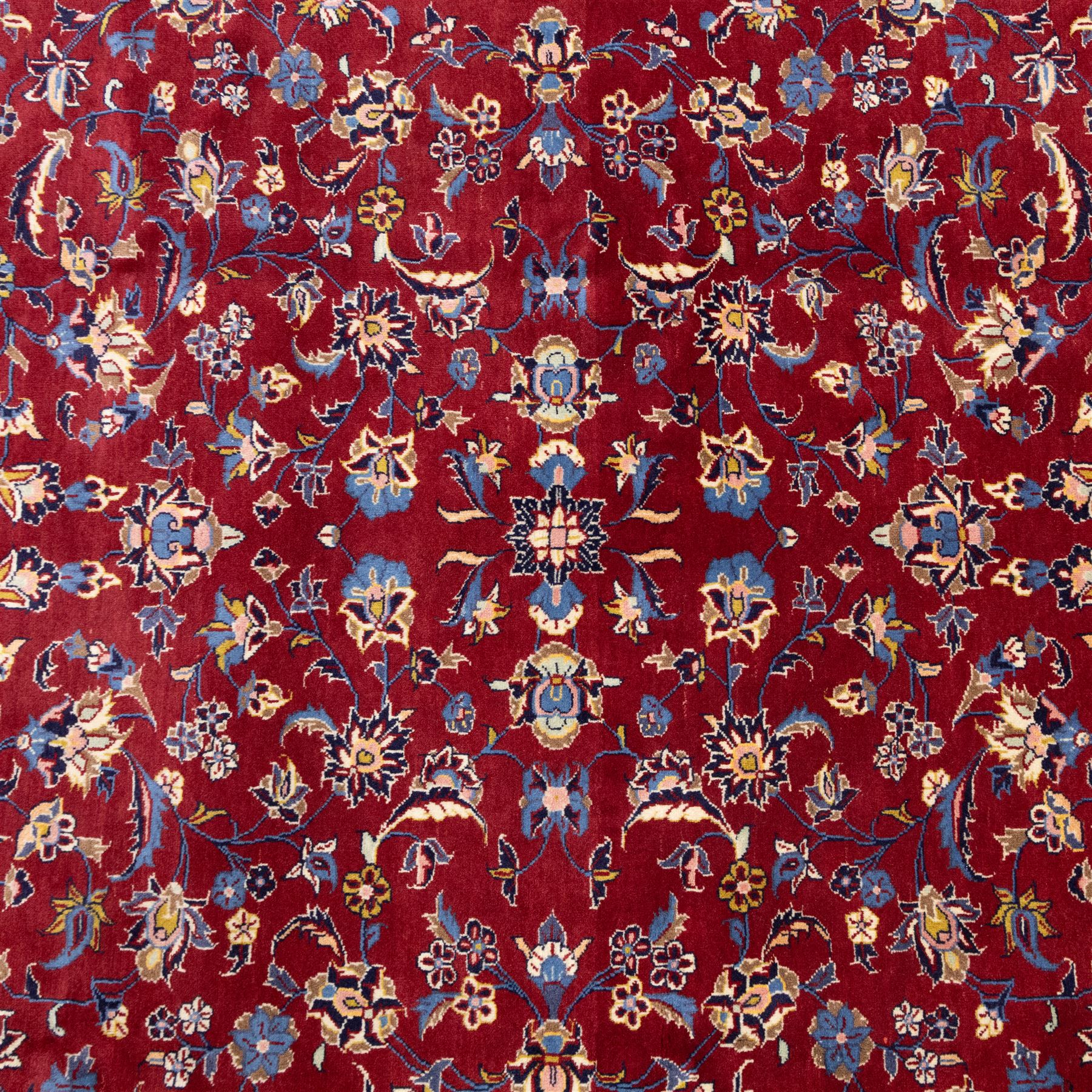 Central Persian Kashan hand-knotted rug, crimson field, decorated with an all-over floral and foliate design incorporating stylized palmettes, rosettes, and scrolling vine work, enclosed by a dark navy blue main border ornamented with flowering branches and palmettes flanked by multiple narrow guard borders with repeating floral motifs, with cotton fringe ends 