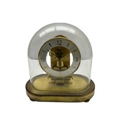 A  late 20th century German Kieninger & Obergfell, “Kundo” battery operated 
mantle clock under an acrylic shade, with an electrically operated solenoid pendulum on an oval brass base with three adjustable feet, visible escapement through an open chapter ring dial, with gilt hands, three-hour numerals and baton markers, with pendulum lock.  H23cm W23cm D13cm
