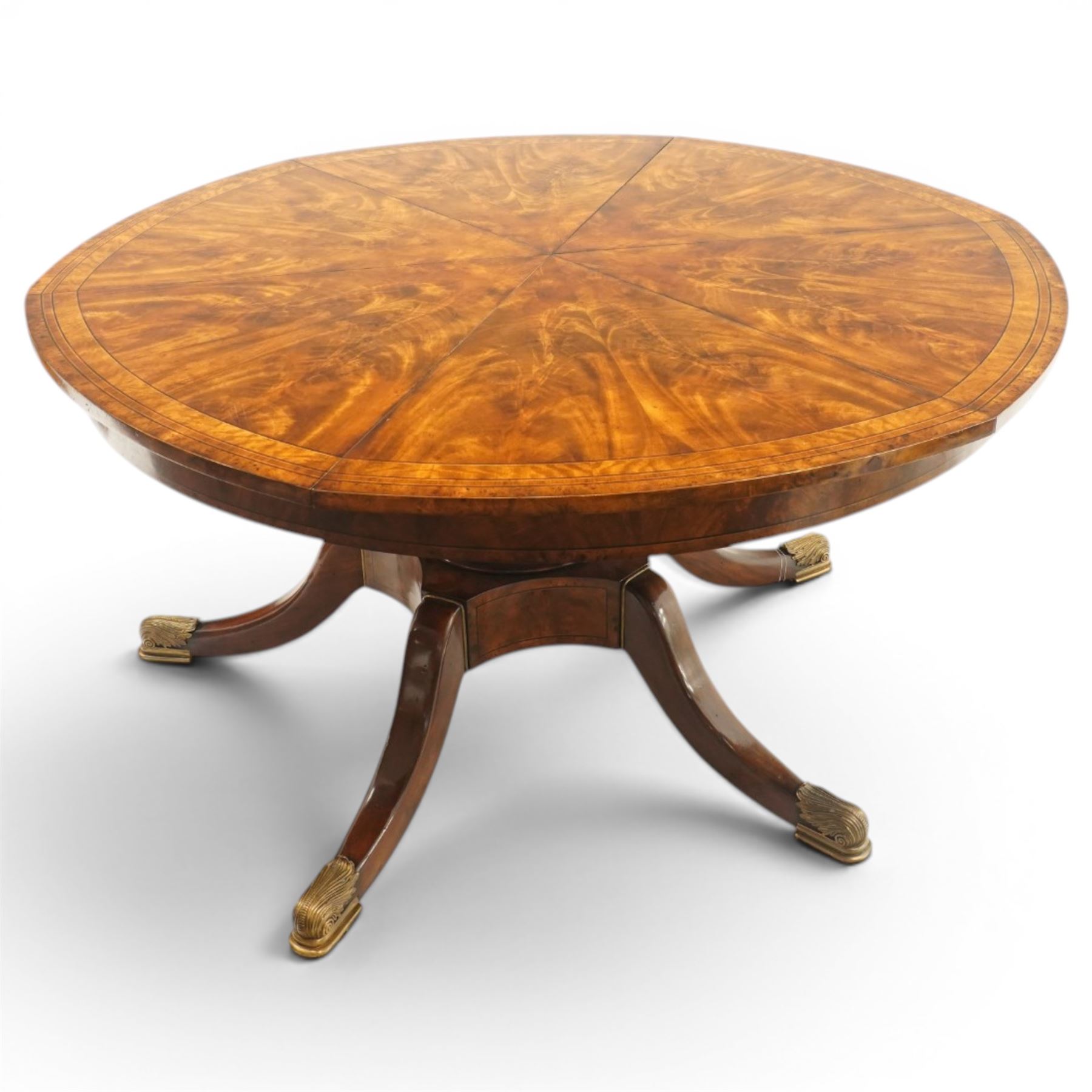 Theodore Alexander - Regency Revival figured mahogany 'Althorp Patent Jupe' extending dining table, the segmented circular top formed of six pull-out triangular divisions with inset fold-out leaves, raised on a large turned vasiform pedestal on a pentagonal platform base with ebonised stringing, terminating to five splayed supports with gilt metal acanthus scrolled feet, retailed by David Hick Interiors, Jersey
