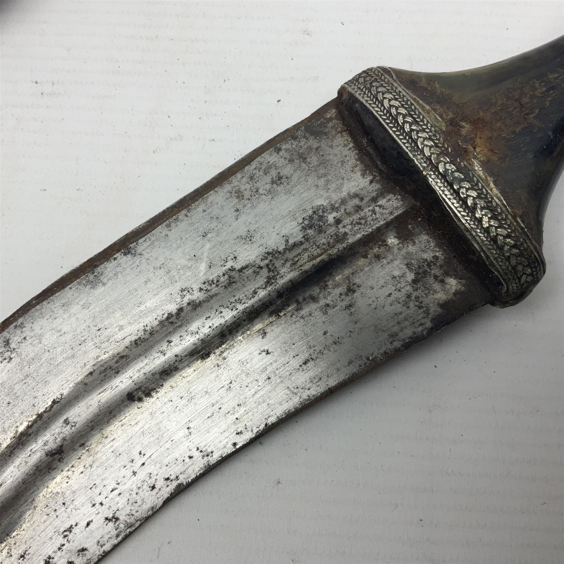 Eastern Jambiya, the 19cm double edge steel blade with raised medial ridge, waisted horn grip with pique work and white metal band; in stitched leather scabbard L33cm overall
