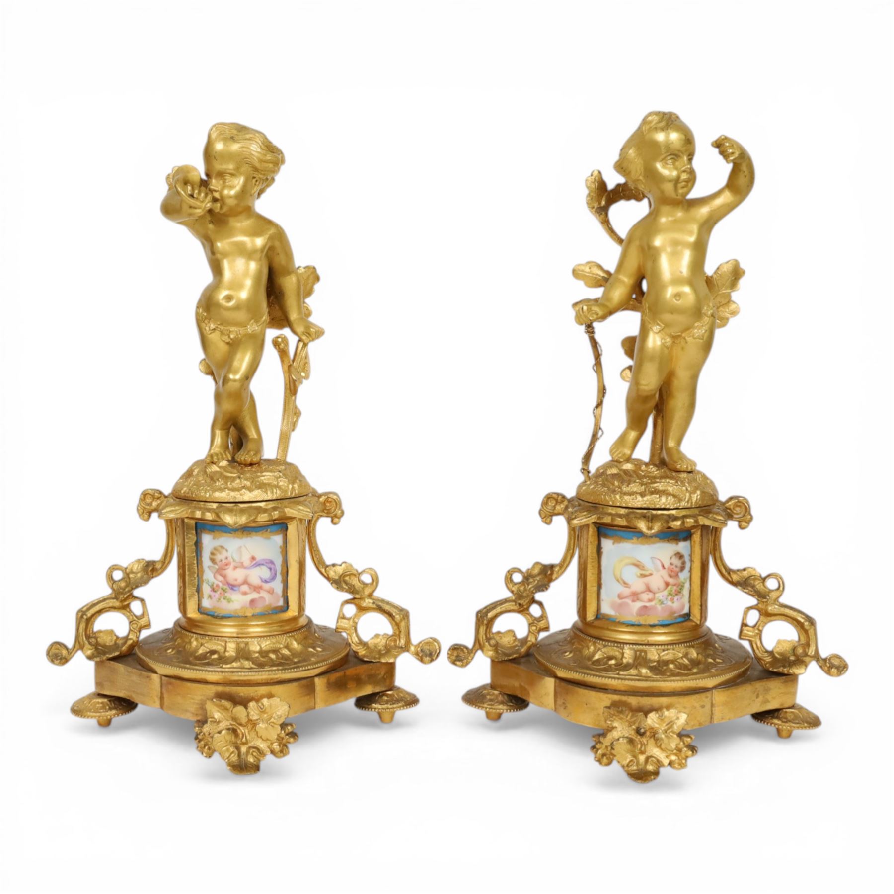Pair of 19th century ormolu putto figures, each modelled stood with foliate support to backs, upon a naturalistic base upon cylindrical plinth each with twin leafy tendril supports, each plinth with inset Sevres type painted panel of a cherub, upon a shaped base with four compressed disk feet, the front facing with leaf mount, H29cm