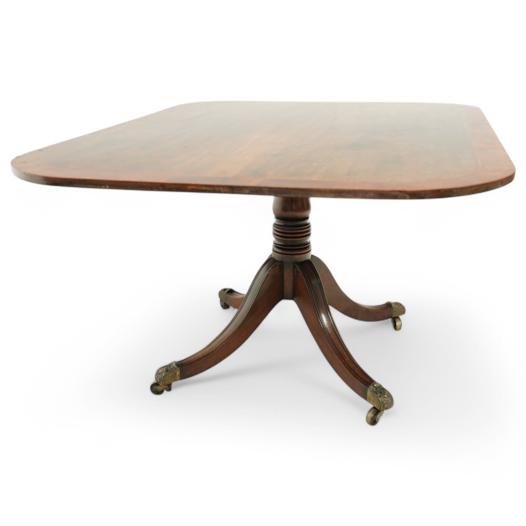 Early 19th century Sheraton style mahogany and rosewood pedestal breakfast table, the rectangular top with rounded corners and crossbanded edge, raised on a turned column support with reeded detail, terminating in four splayed legs with brass caps and castors