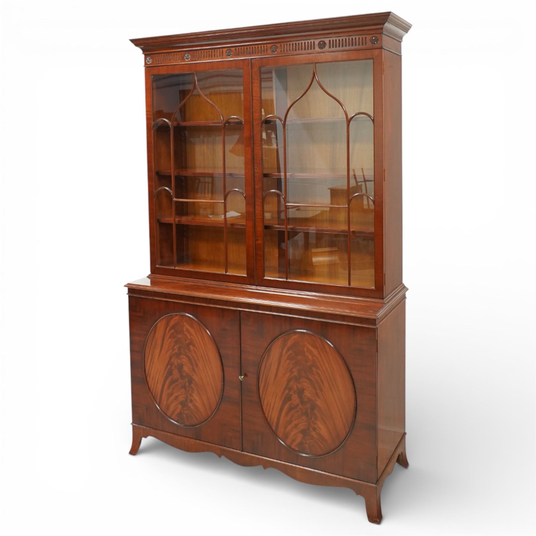 William Tillman - Early 20th century mahogany display cabinet, moulded cornice above a frieze carved with roundels and fluting, pair of astragal glazed doors enclosing adjustable shelving, the lower section fitted with twin panelled cupboard doors centred with oval veneered panels, shaped apron and bracket feet
