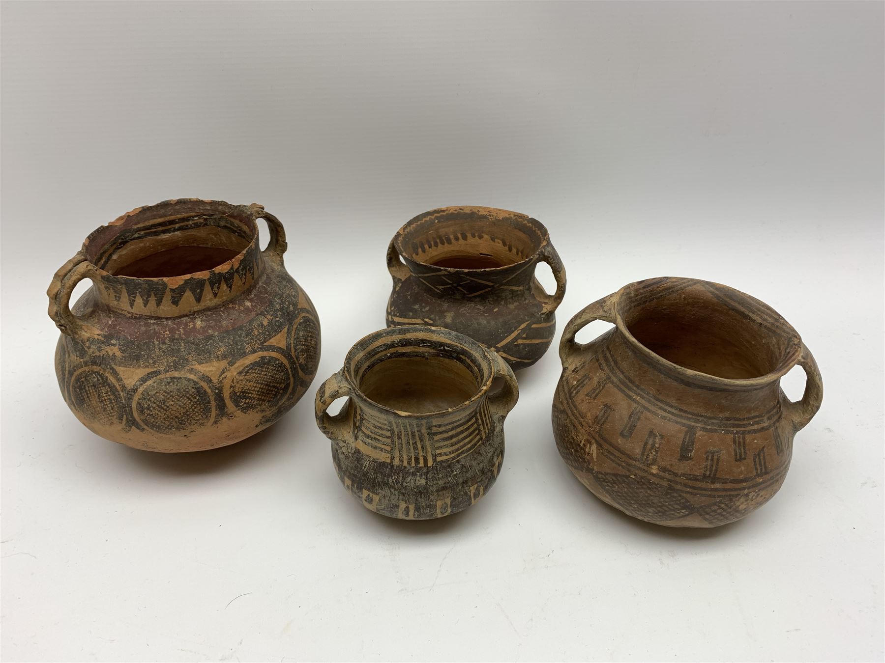Four Chinese Neolithic pottery vessels, each with twin loop handles to the shoulder and painted geometric banded decoration to the body, three bearing labels for Eskenazi London, tallest H13cm
