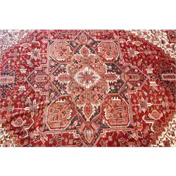 North West Persian Heriz hand-knotted carpet, brick red field with a large central angular star medallion with pendants surrounded by stylized geometric palmettes, serrated leaves and tribal motifs,, ivory corner spandrels, field enclosed by a dark indigo main border of stylize rosettes and angular vine motifs between multiple guard stripes, with cotton fringe ends 