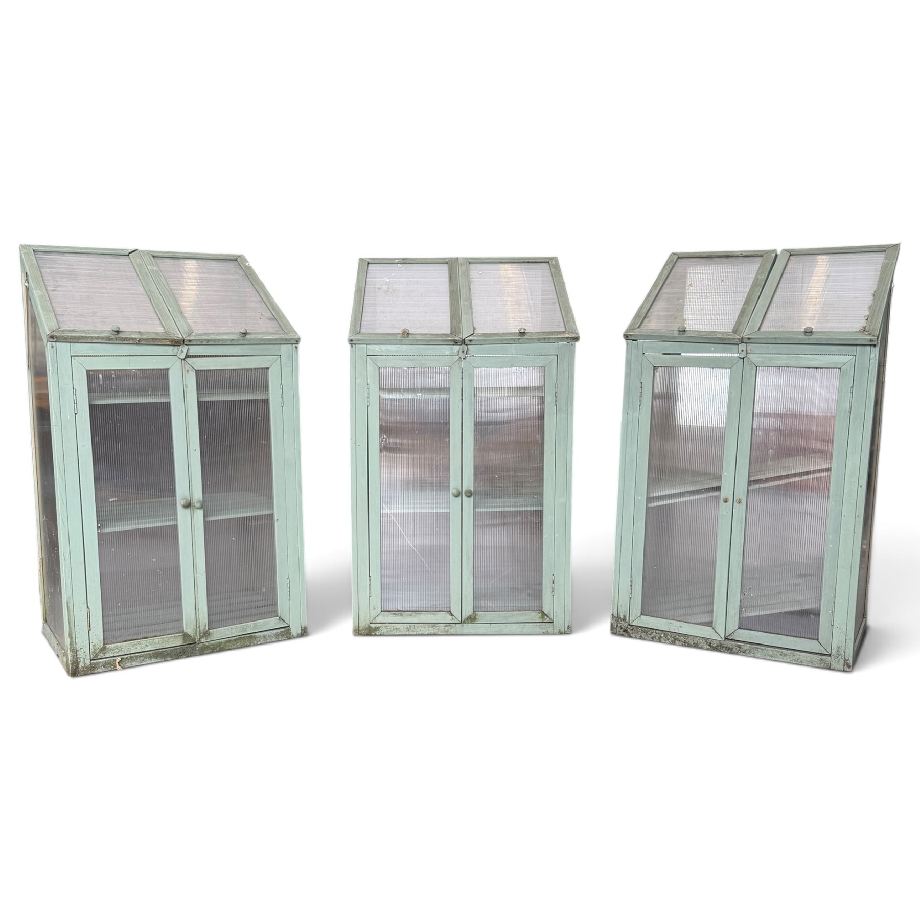 Three green-painted timber cold-frame greenhouses or growhouses, each with twin hinged sloping polycarbonate lids above a pair of panelled doors enclosing slatted shelves