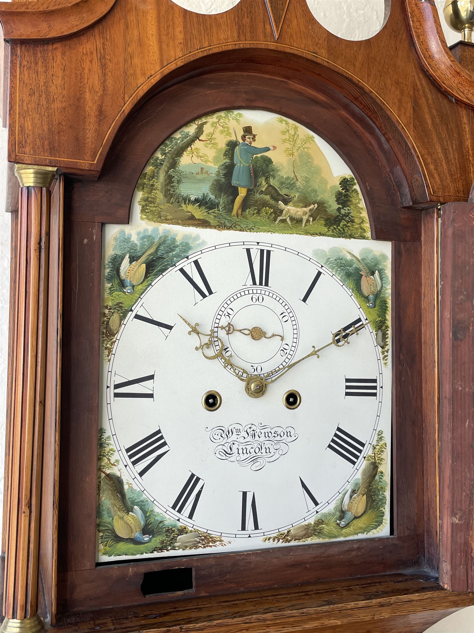 An oak and mahogany longcase clock by William Hewson, Lincoln c 1860, with a swans neck pediment, ball and spire finials and brass paterae, break arch hood door flanked by reeded pilasters with brass capitals, oak trunk with canted corners and a short oak door with mahogany crossbanding, conforming plinth with applied skirting, fully painted dial with Roman numerals, minute track and subsidiary seconds dial, matching brass stamped hands and winding collets, depiction of a  sportsman to the break arch and corresponding game birds to the spandrels, dial pinned directly to an eight-day rack striking movement with a recoil anchor escapement, striking the hours on a bell. Original casemakers label pasted to the inside of the case 