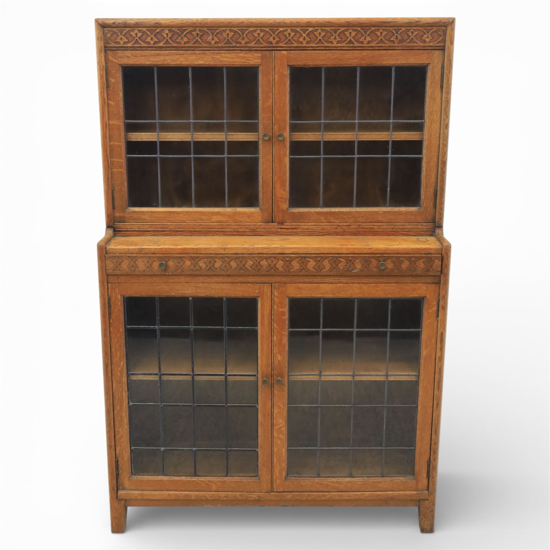 Early 20th-century Arts and Crafts glazed oak bookcase, upper section fitted with a pair of leaded glazed doors enclosing a fixed shelf above a projecting frieze drawer interior with baize-lined writing surface flanked by two compartments, carved with interlaced decoration, lower section fitted with a further pair of leaded glazed doors enclosing a single shelf, raised on short stile feet 