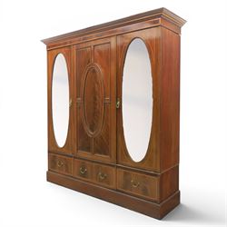 Edwardian inlaid mahogany triple wardrobe, featuring two oval mirror doors with bevelled plates, enclosing central figured veneered panel with mouled detail, above three base drawers