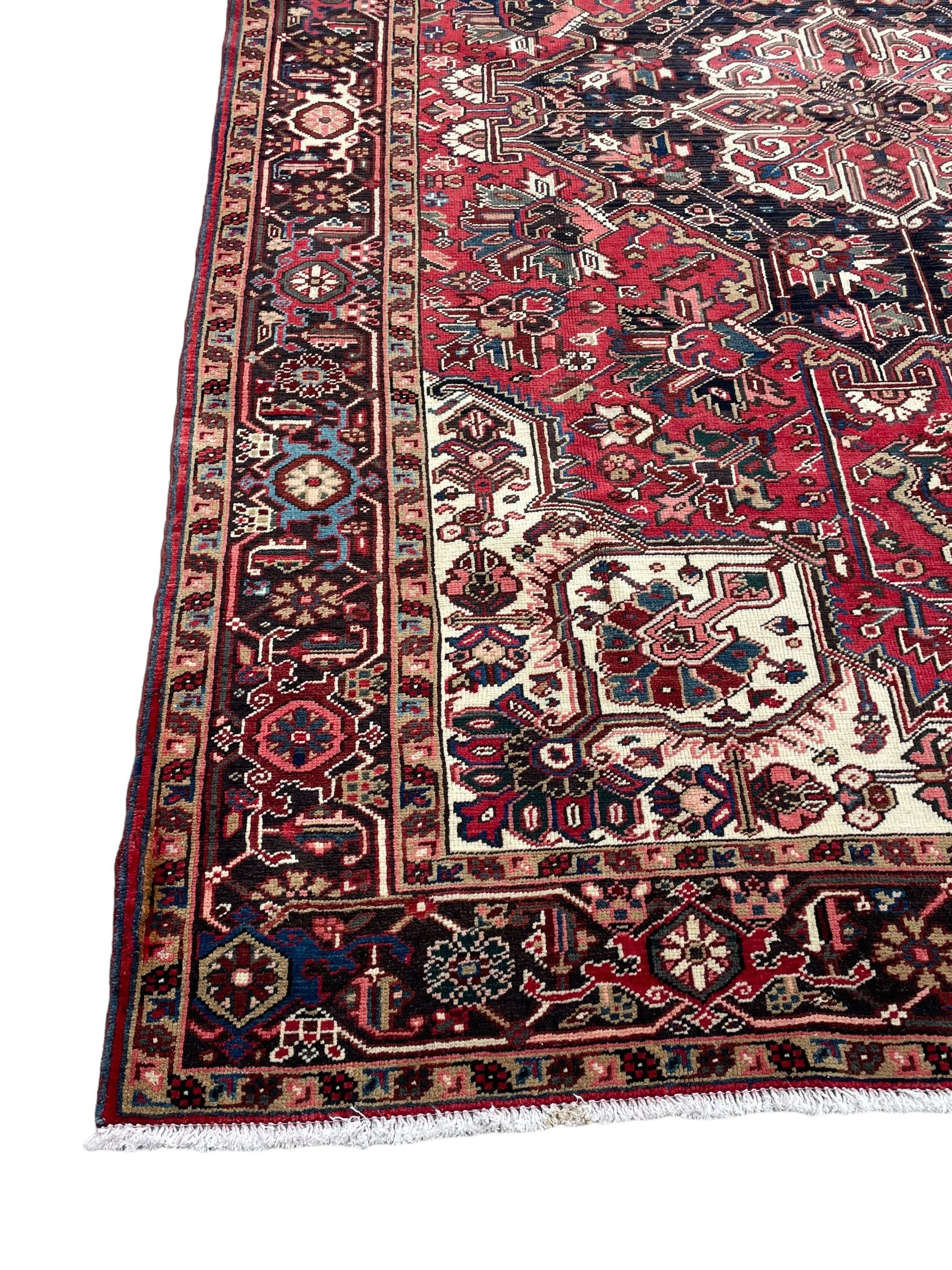 Persian Heriz red ground rug, central scrolled medallion on dark ground decorated with floral motifs, the surrounding field decorated with stylised plant motifs and smaller geometric symbols, guarded border with repeating pattern punctuated with rosettes and flower heads 