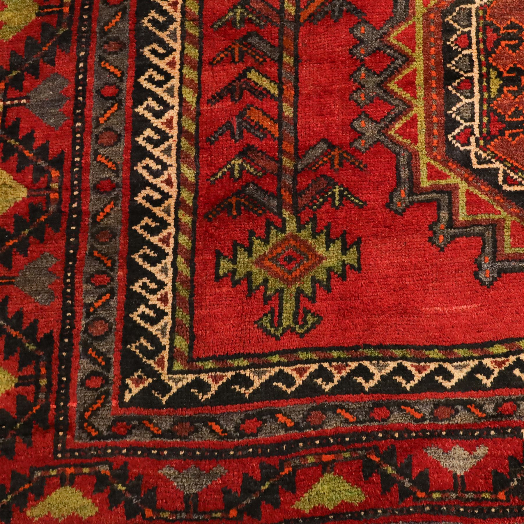 North East Persian Kurdish hand-knotted carpet, madder red field with three vertical and central stepped octagonal medallions enclosing angular geometric motifs with scattered tribal devices, framed by multiple narrow borders with latch-hook patterns, enclosing a bold main border of repeating geometric motifs, palette with indigo, dark brown and soft green tones, 