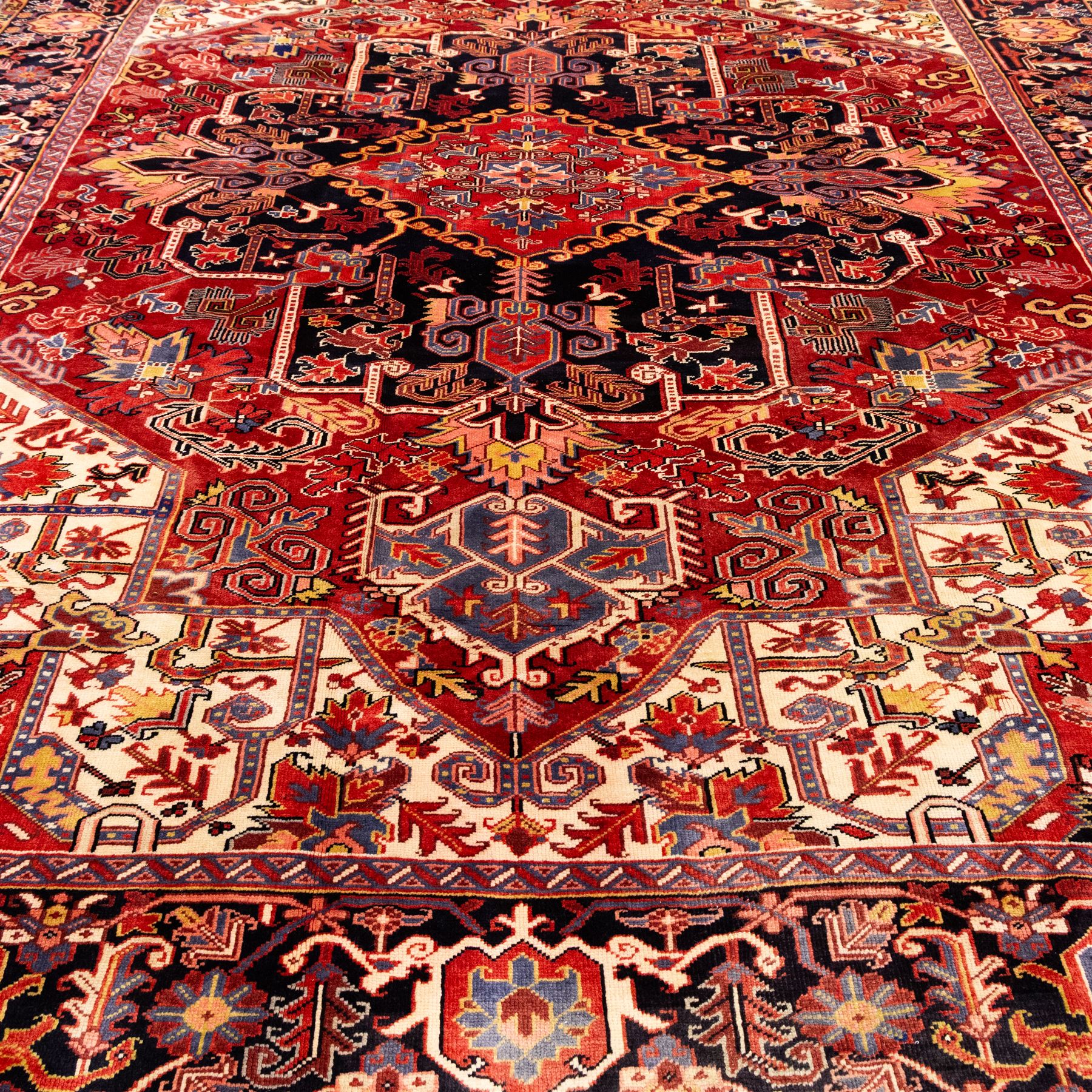 North West Persian Heriz hand-knotted carpet, madder red field with large central stepped medallion with angular floral and geometric designs, surrounded by stylized palmettes, leaves and tribal motifs with indigo corner spandrels, enclosed by a broad navy border of geometric floral devices between multiple guard stripes, with cotton fringe ends 