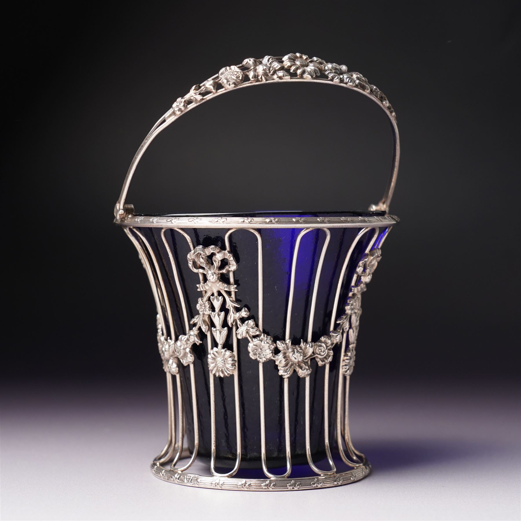 Edwardian pierced silver sugar basket with swing handle and blue glass liner H9cm London 1906 Maker Haseler Bros. and another decorated with floral swags Chester 1912 Maker  (2)