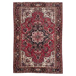 Persian Heriz red ground rug, central scrolled medallion on dark ground decorated with floral motifs, the surrounding field decorated with stylised plant motifs and smaller geometric symbols, guarded border with repeating pattern punctuated with rosettes and flower heads 
