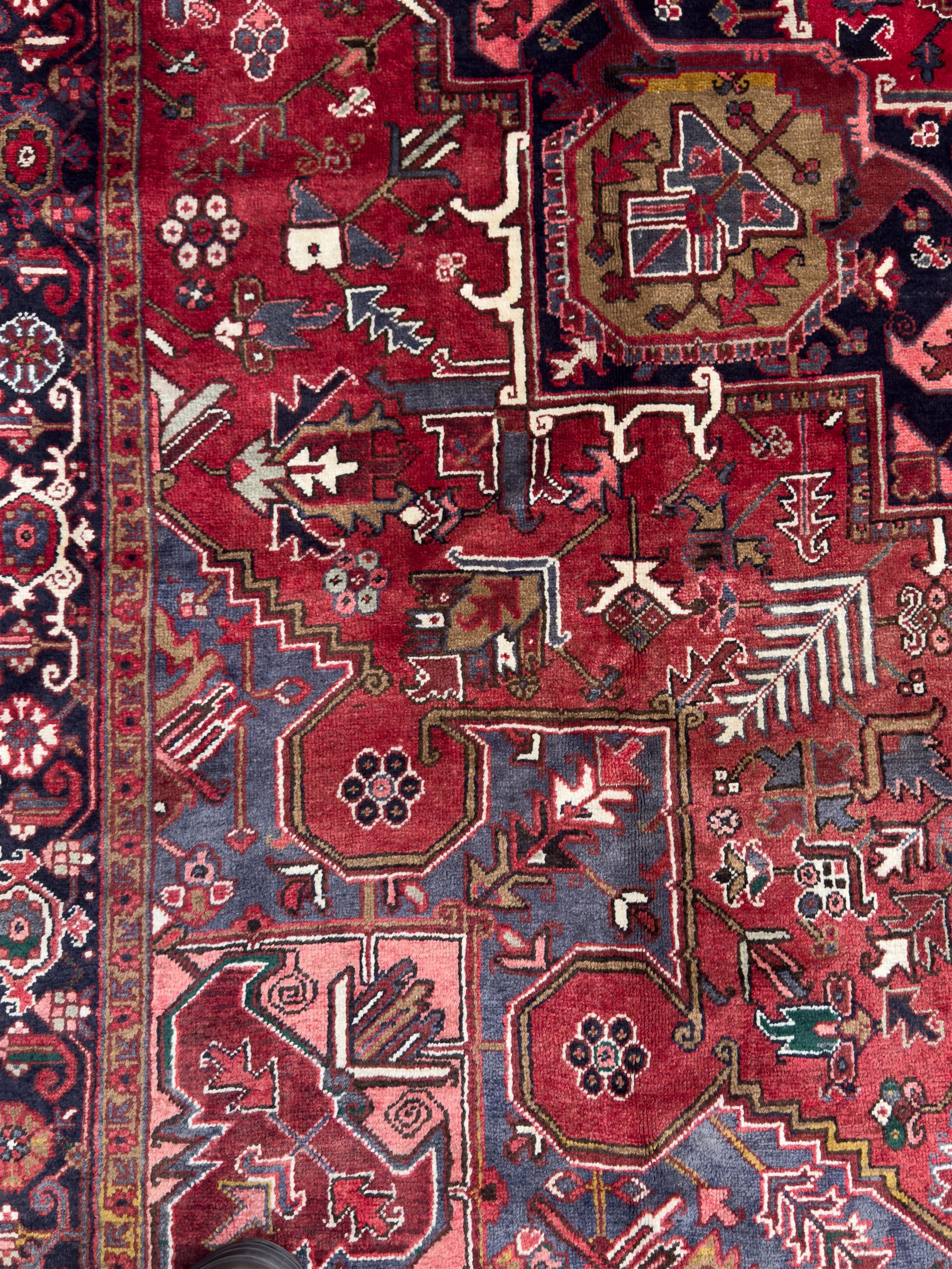 Northwestern Persian Heriz hand-knotted carpet, madder red field with a bold stepped medallion in indigo, ivory, and ivory, surrounded by angular floral motifs and stylized leaves, and the spandrels set within a dark border of rosettes and angular vine motifs, 335cmx252cm
