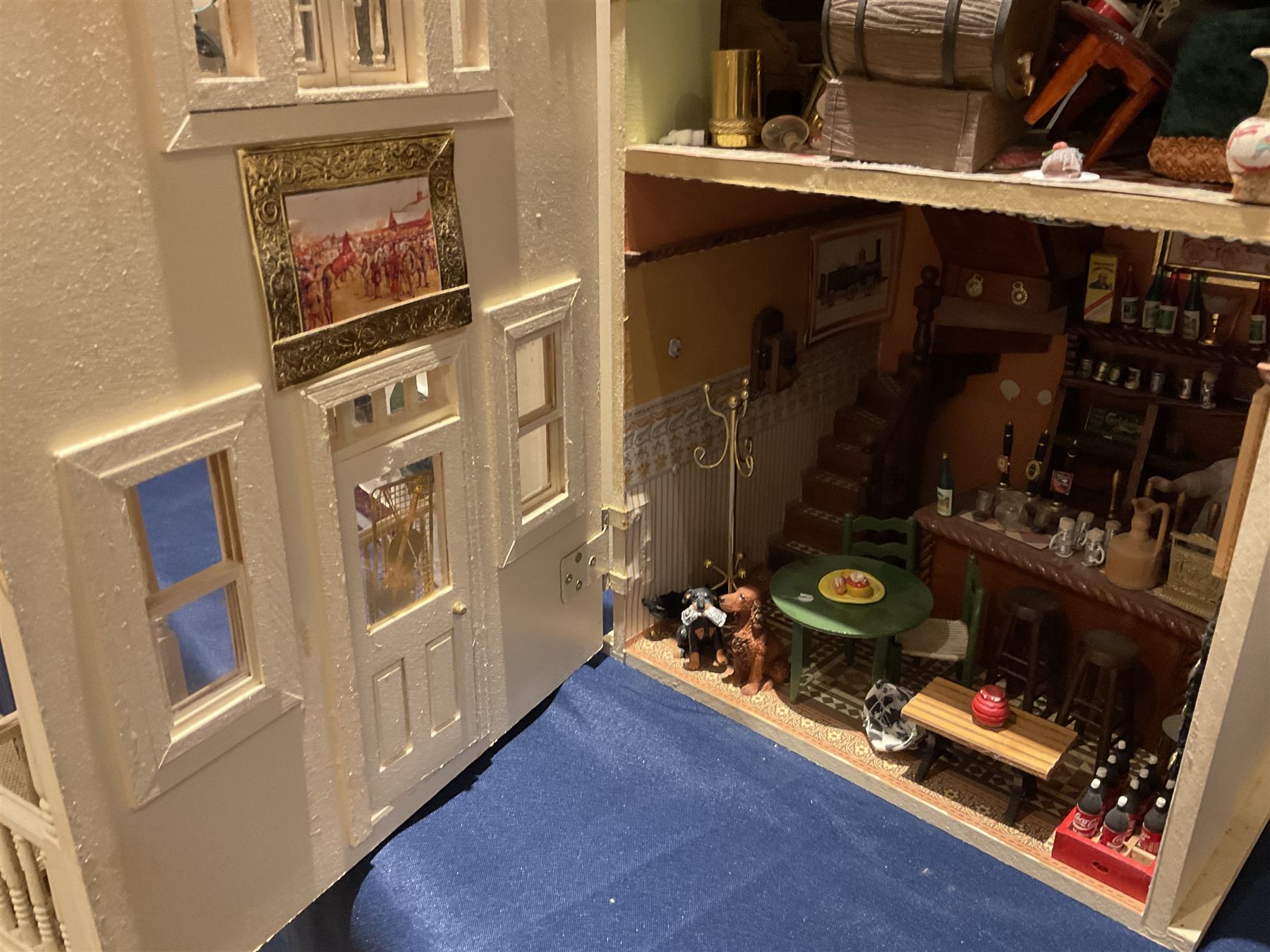 Seagull Pub - scratch-built wooden doll's house as a two-storey gable fronted public house painted in white green and maroon; the hinged front with ground floor veranda and window over opening to reveal two fully furnished rooms with dog-leg staircase and well stocked bar with bar-tender L31cm H62cm D37cm; together with Corner Shop and Dollies Pantry; a similarly painted scratch-built wooden two-storey flat-roofed doll's house as a well stocked ground floor grocery shop with stairs up to two further fully furnished rooms and more stairs giving access to a roof terrace L46cm H57cm D33cm (2)