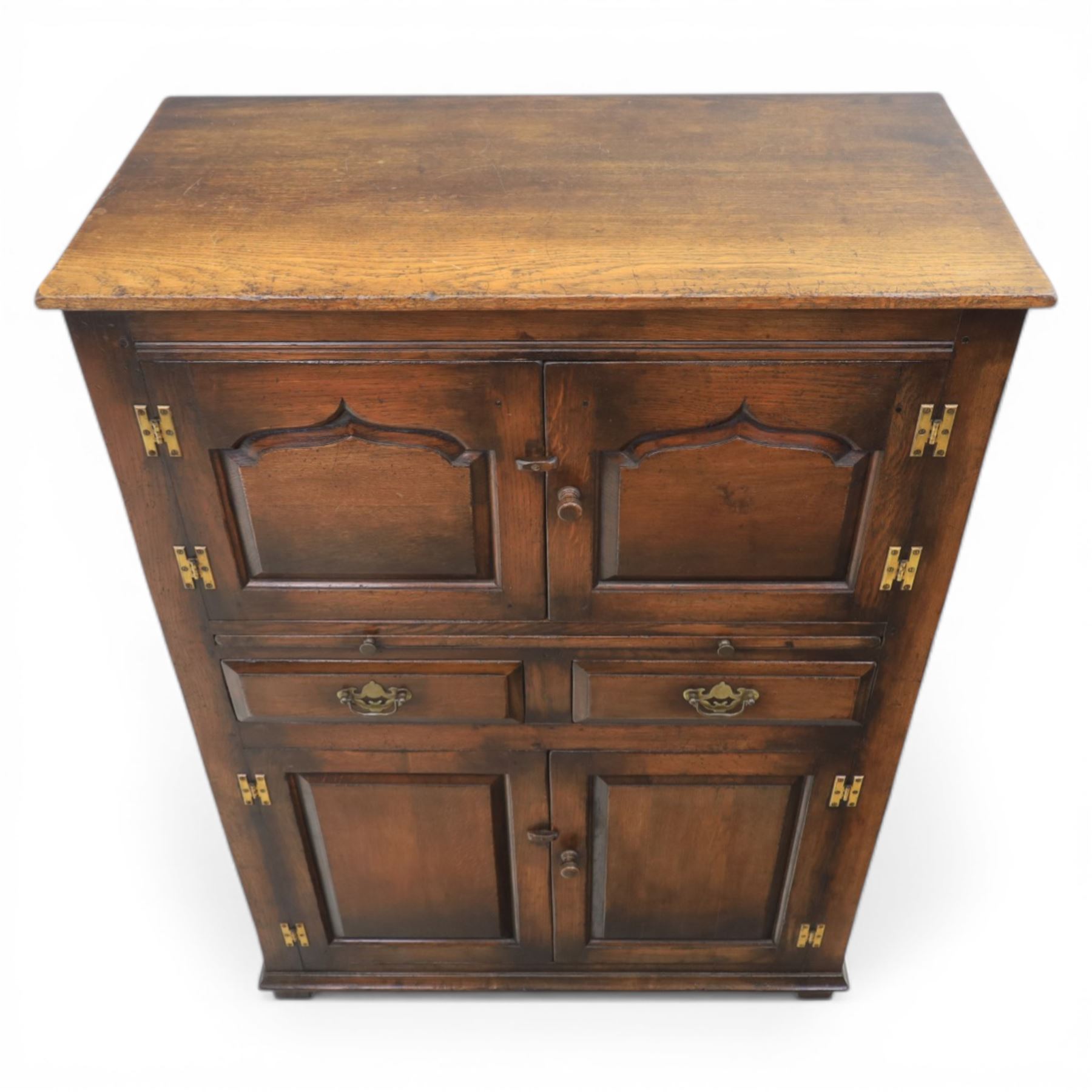  Late 20th century oak four door cupboard in the manner of Titchmarsh and Goodwin, rectangular plank top above two pairs of fielded panel doors with shaped raised panels, central arrangement of a brushing slide above two short drawers with brass swan neck handles and pierced backplates, enclosed by further pair of panelled cupboard doors, fitted with brass butt hinges and turned wooden knobs, raised on plinth base