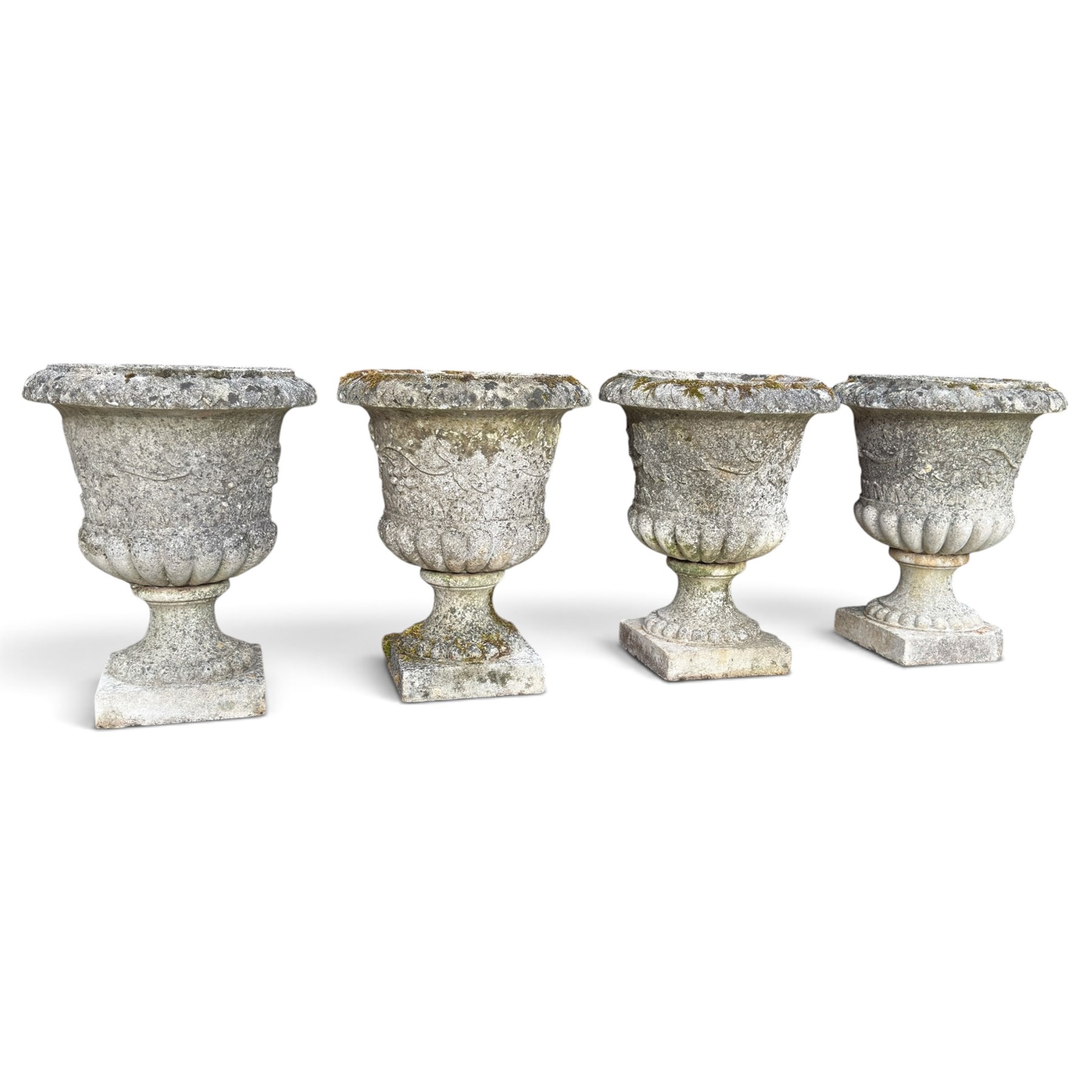 Set of four weathered composite stone urns of campana form, the everted leaf-moulded rolled rim over a body decorated with scrolling fruiting grapevines in relief, over a gadrooned underbelly and waisted socle, lobed collar on square base