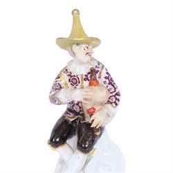 Mid 18th century Meissen figure, modelled as a harlequin playing bagpipes, circa 1745, possibly modelled by J.F.Eberlein after earlier models by J.J.Kändler, wearing a gilt edged tunic painted with flowers, black breeches with rosettes at the knees, red shoes and conical yellow hat, seated upon a rock, with traces of blue crossed swords mark beneath, H14cm