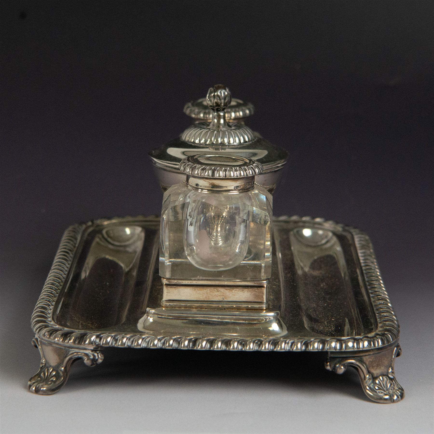George IV silver desk stand, of rectangular form with rounded corners and oblique gadrooned rim, upon four embossed pad feet, with central pedestal dish of part fluted form with domed lid engraved with crest and with bud finial, with two recesses either side housing faceted glass inkwells, each with silver collars, hallmarked John & Thomas Settle, Sheffield 1825 & 1827, H11cm