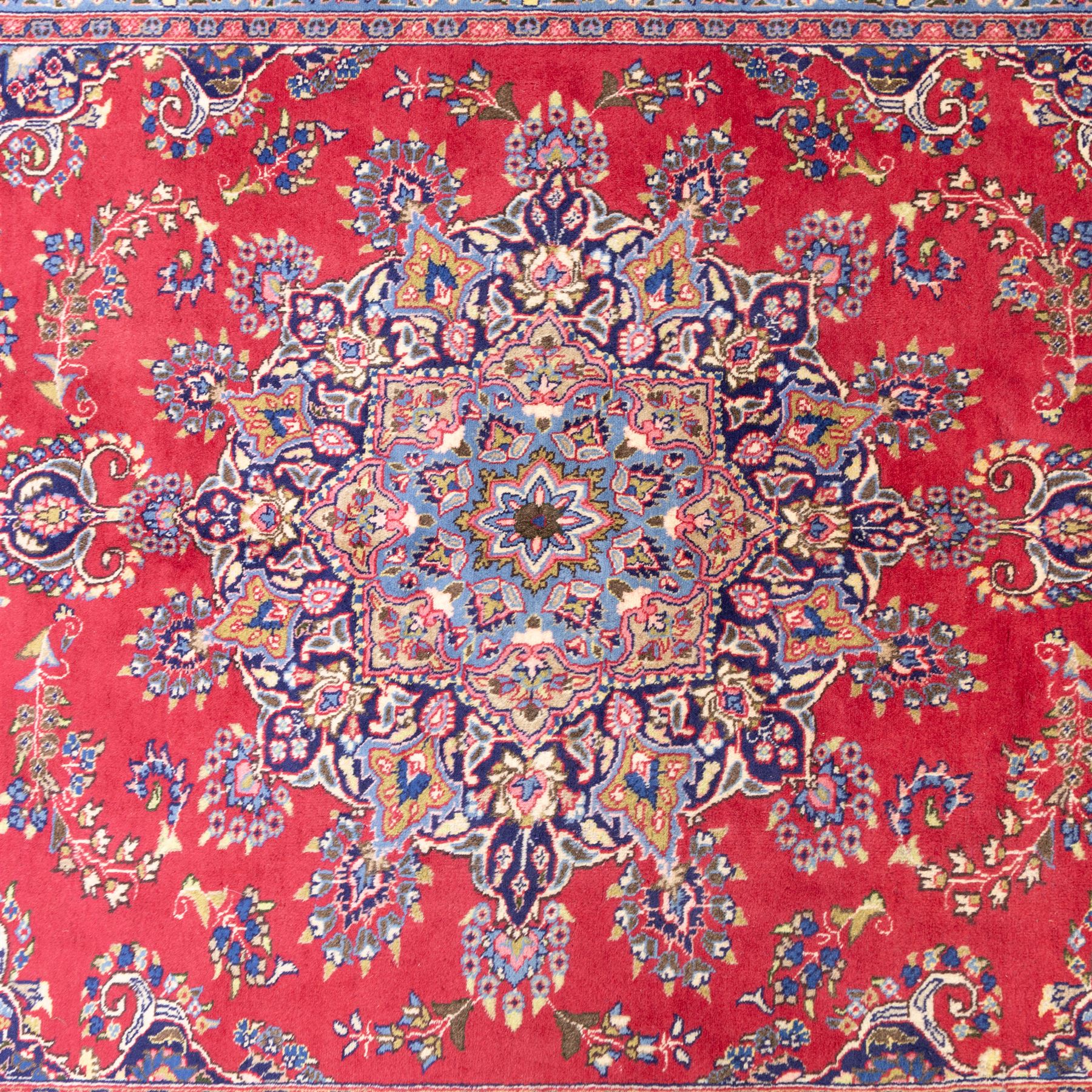 North East Persian Meshed hand-knotted rug, madder red field with central multi-lobbed medallion in shades of indigo, ivory, and sofa pastel tones surrounded by scrolling floral vines, palmettes, stylized blossoms, floral sprays, and cartouches, framed by Indigo spandrels, enclosed within a broad dark blue main border, flanked by multiple narrow guard stripes, with fringe ends 
