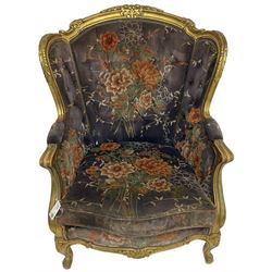 Pair of Louis XVI design gilt framed wingback armchairs, cresting rail carved and moulded with foliate decoration and C-scrolls, upholstered in buttoned floral patterned mauve velvet with loose seat cushion, scallop carved apron over cabriole supports
