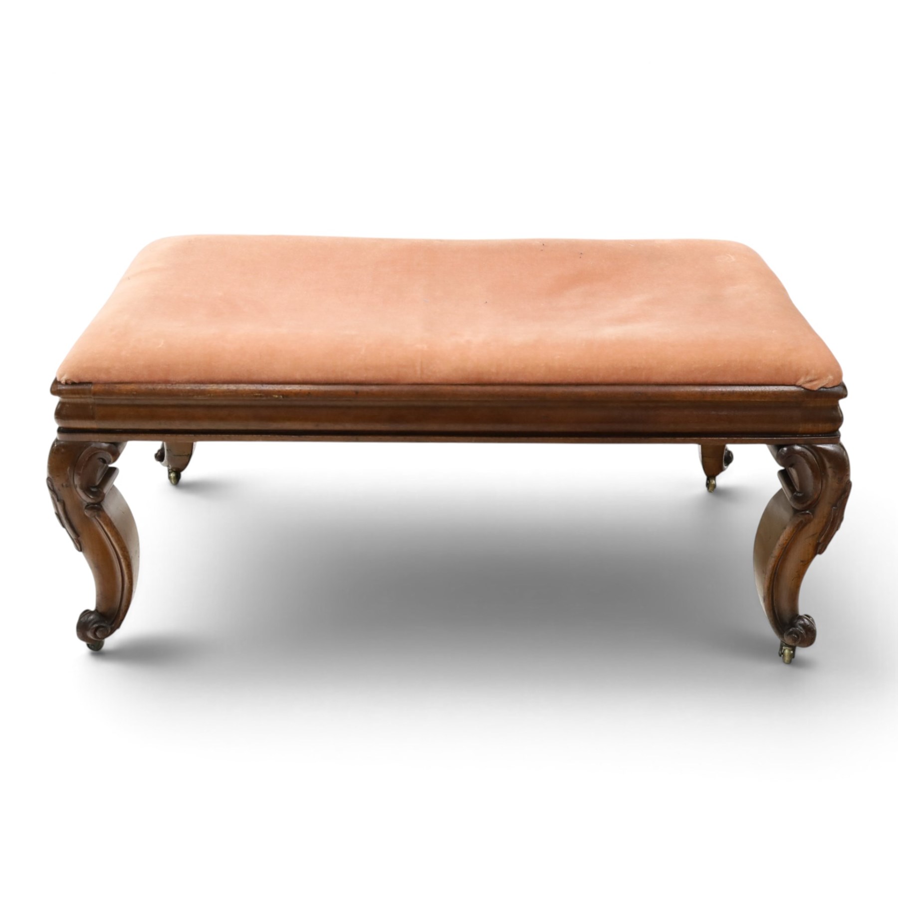 Victorian carved mahogany fender stool of generous proportions, the wide rectangular seat upholstered in rose fabric, shaped frieze rail over C-scroll and cartouche carved cabriole supports with scroll feet and brass castors