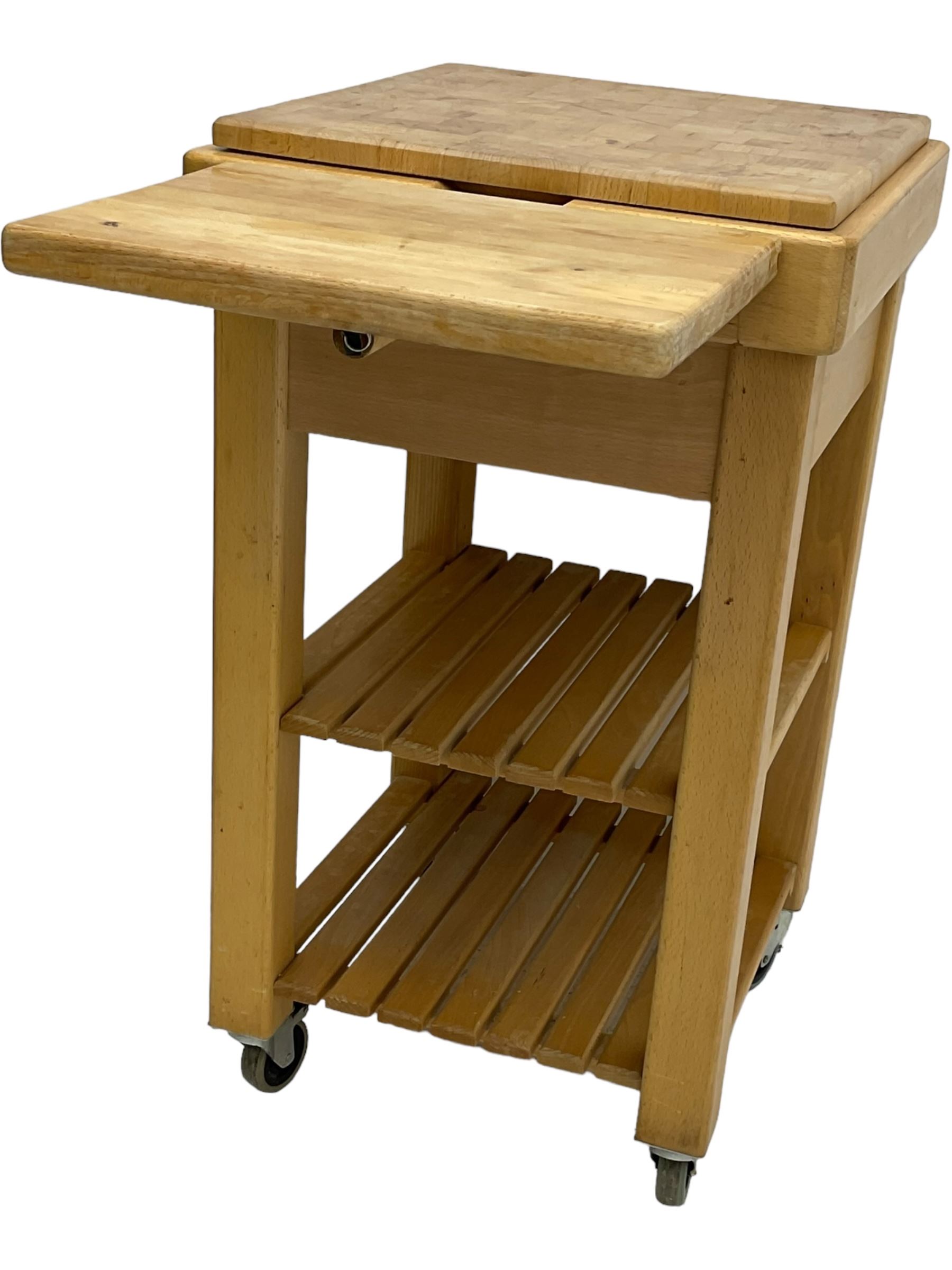 Beech butcher block kitchen cart, featuring square butcher block top, side drop-leaf extension, single drawer with metal sides, and two slatted shelves below, the cart supported by four castors