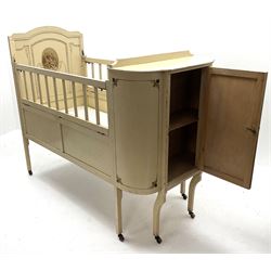 Early 20th century child’s cot, cream painted with applied oval putti scenes, the curved end fitted with cupboard, drop down sides, on square supports with castors