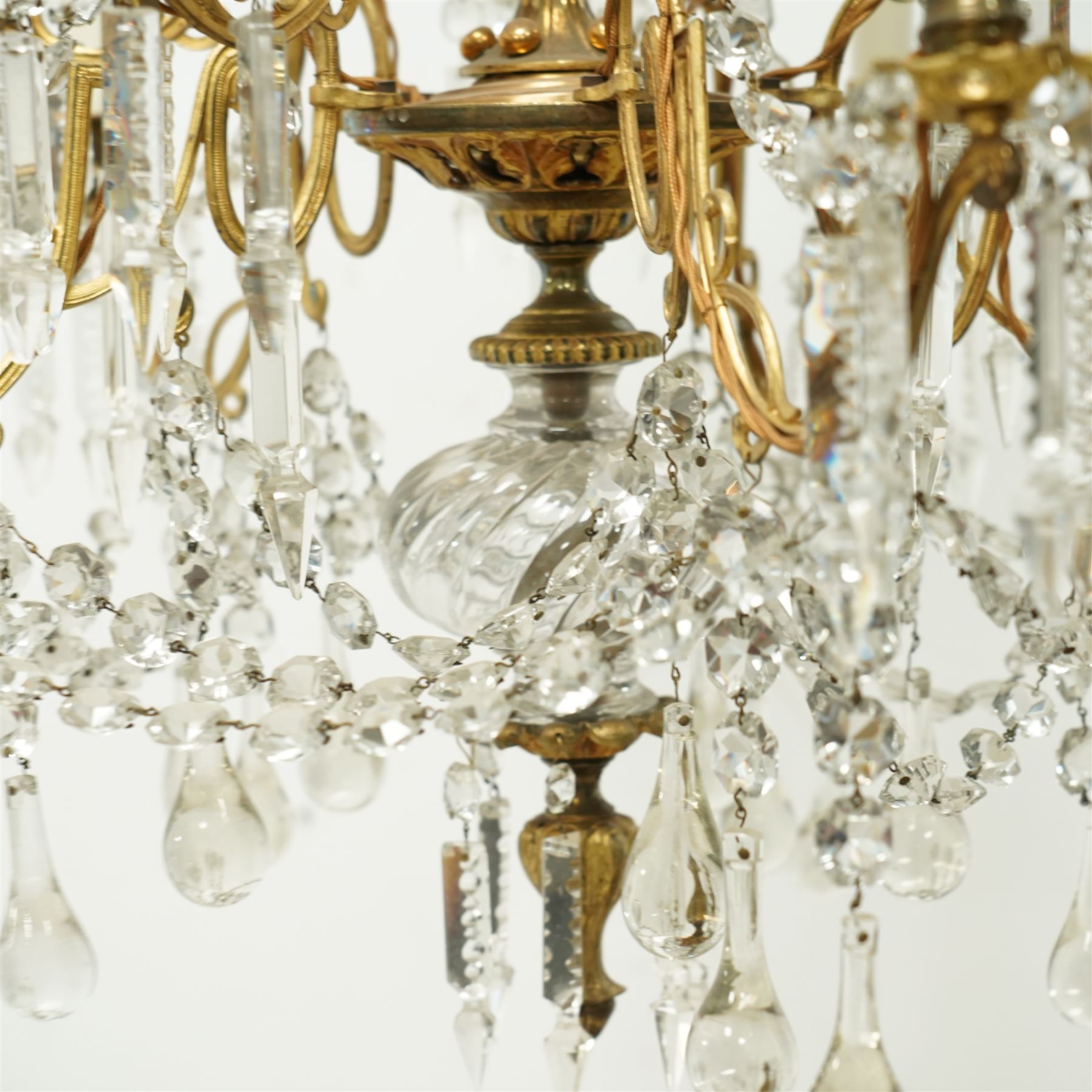 Late Victorian gilt bronze mounted crystal chandelier, ceiling rose above shaped top of trailing swags, glass crystal droplets, beneath rising curled branches and glass beads tapering down to five candle sconces, inside circular bezel fitted with drops and glass twist fluted stem, supported with an internal metal shaft above a rococo style stem holding double branches, and similar styled branches, with twelve branches fitted with candle holders and drip trays with crystal drops and cut crystal spears, the underside festooned with crystal glass swags and a bulbous fluted glass terminal and shaped finial with crystal glass swags attached to the branches, the base with pear shaped droplet graduating to the centre, wired for electricity 