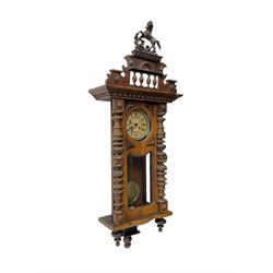 1890’s German striking wall clock in a walnut case, striking the hours and half hours on a coiled gong, eight-day Juhngans movement with a two-part dial, Roman numerals, gothic steel hands and visible gridiron pendulum, case with turned pilasters, applied carving and glazed full length door.

